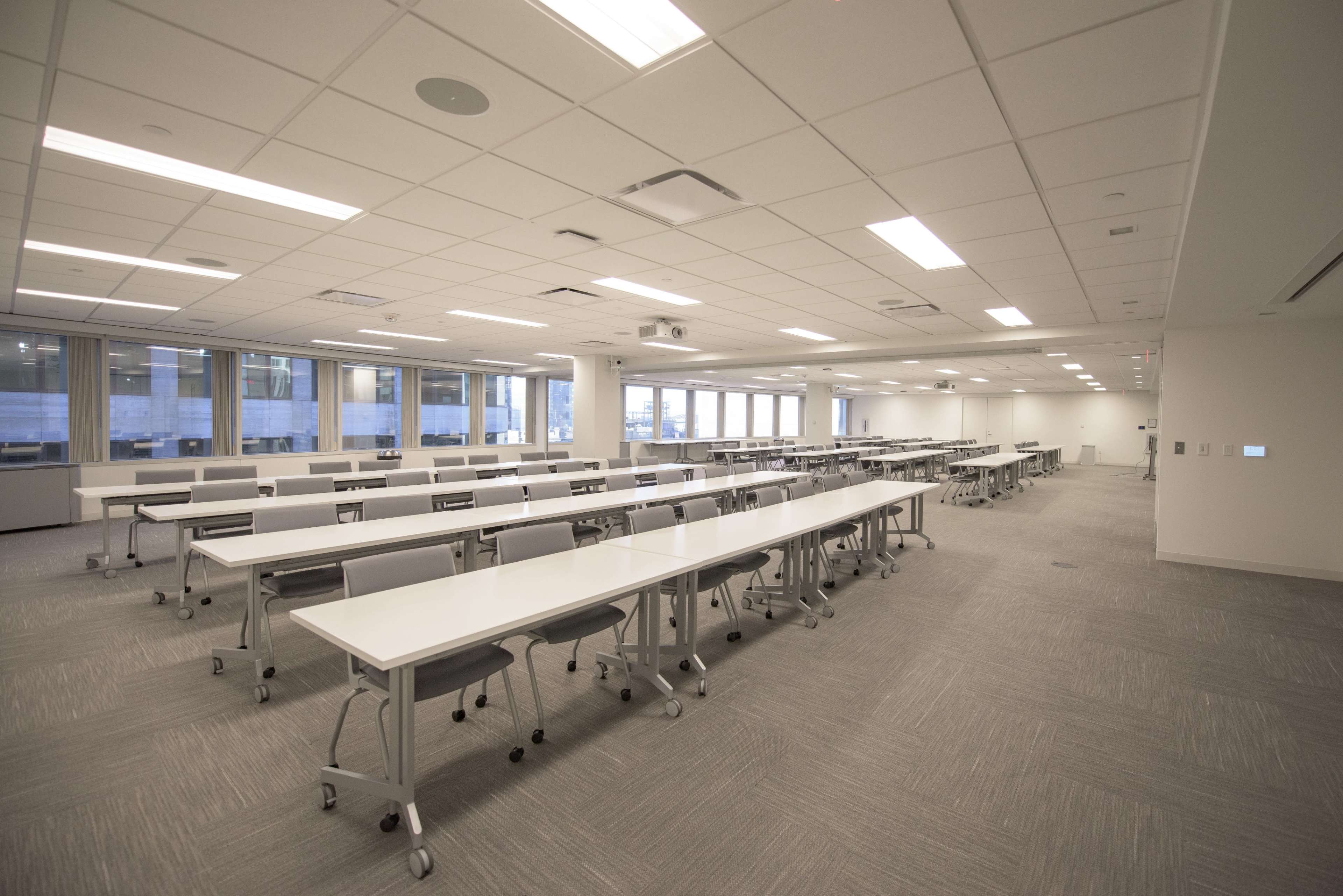 The image shows a spacious, well-lit classroom with multiple long tables and chairs arranged in a grid layout.