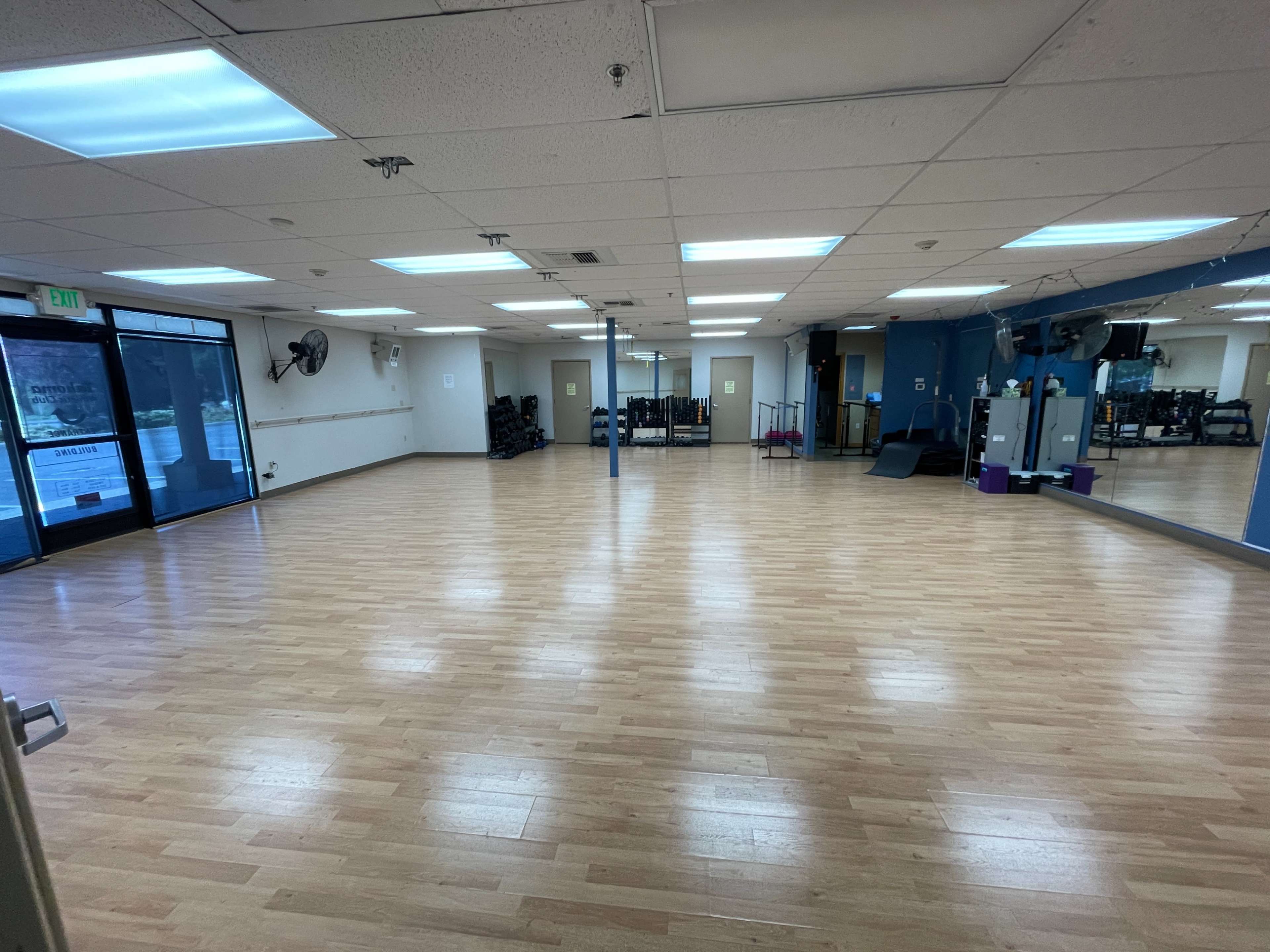 An empty, well-lit fitness studio features wooden flooring, large mirrors, and fitness equipment along the walls.