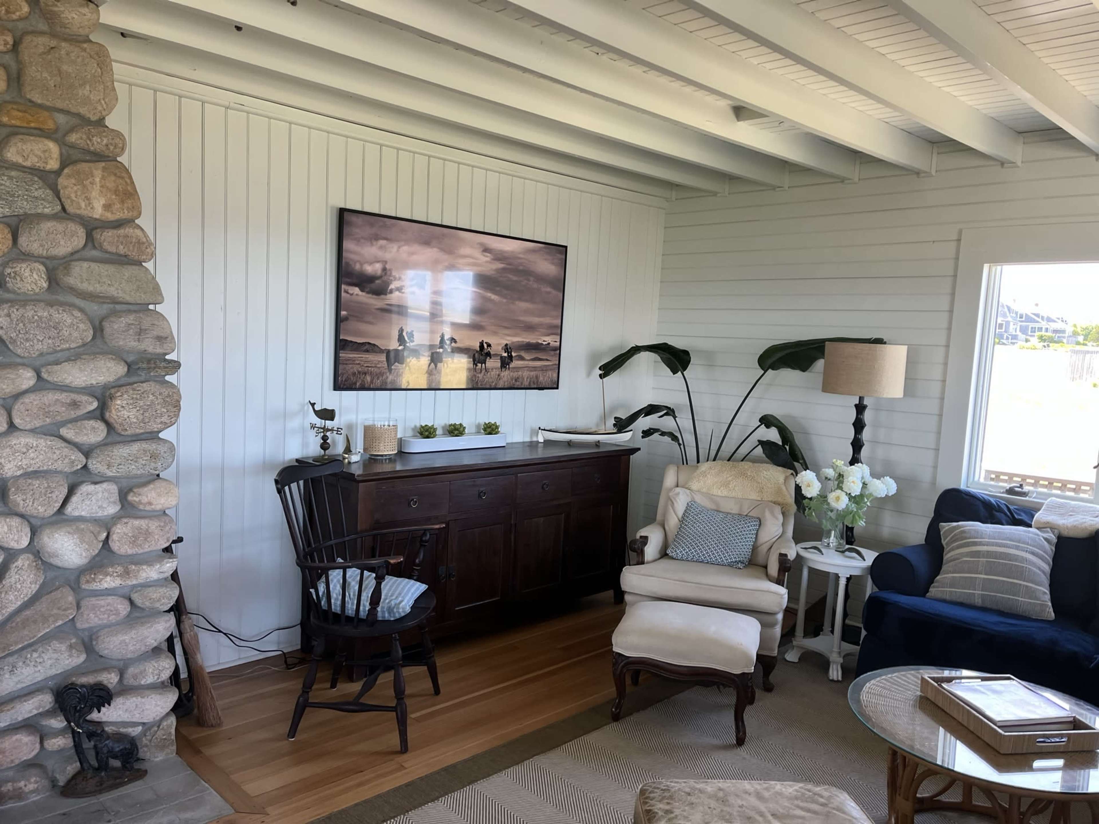 A living room with a stone fireplace, a wall-mounted television displaying a landscape photo, a wooden sideboard, and various seating, including an armchair and a couch.