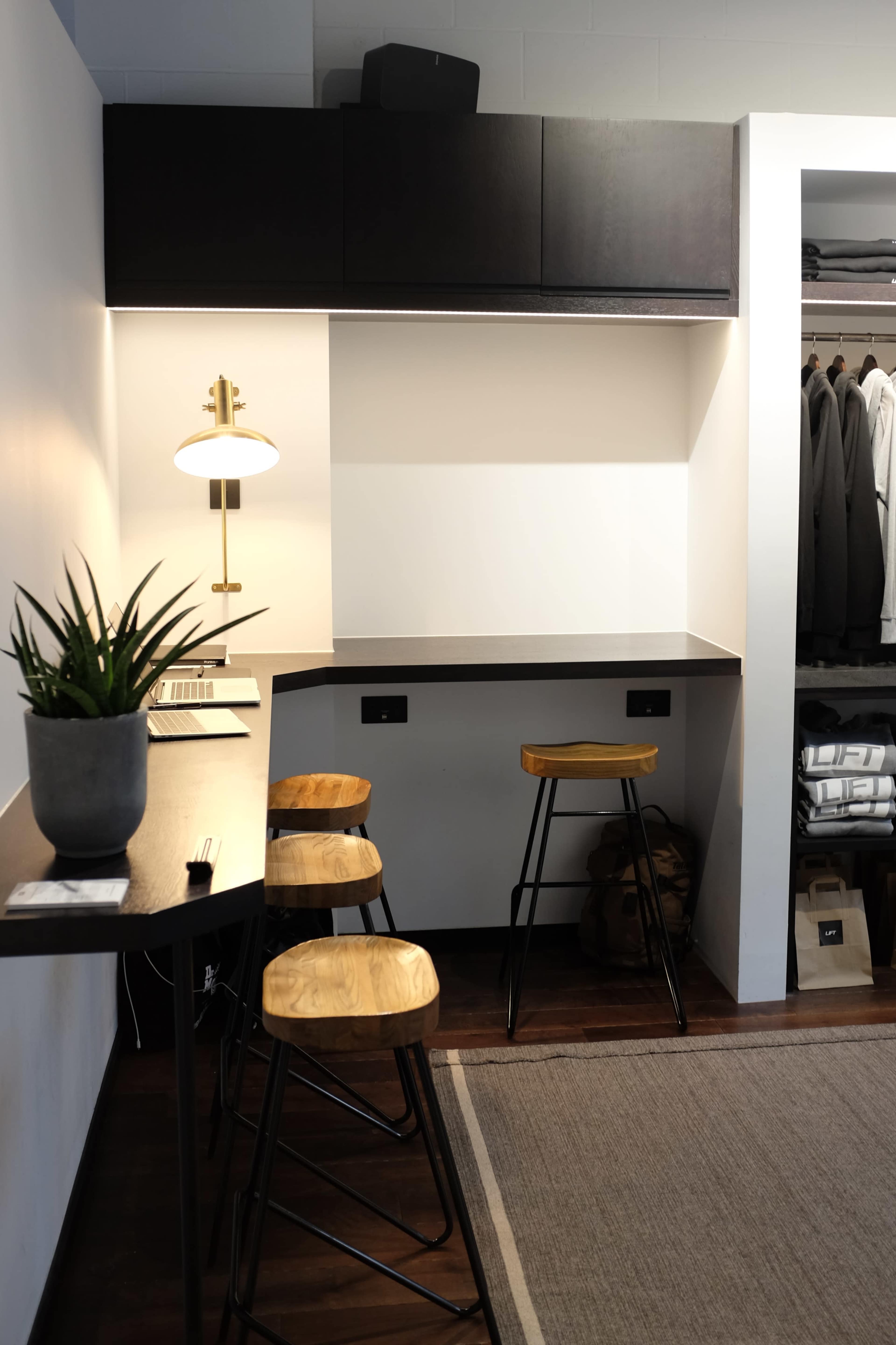 A compact workspace with a wooden table, three metal-framed stools, a potted plant, a lamp, and a neatly arranged closet in the background.