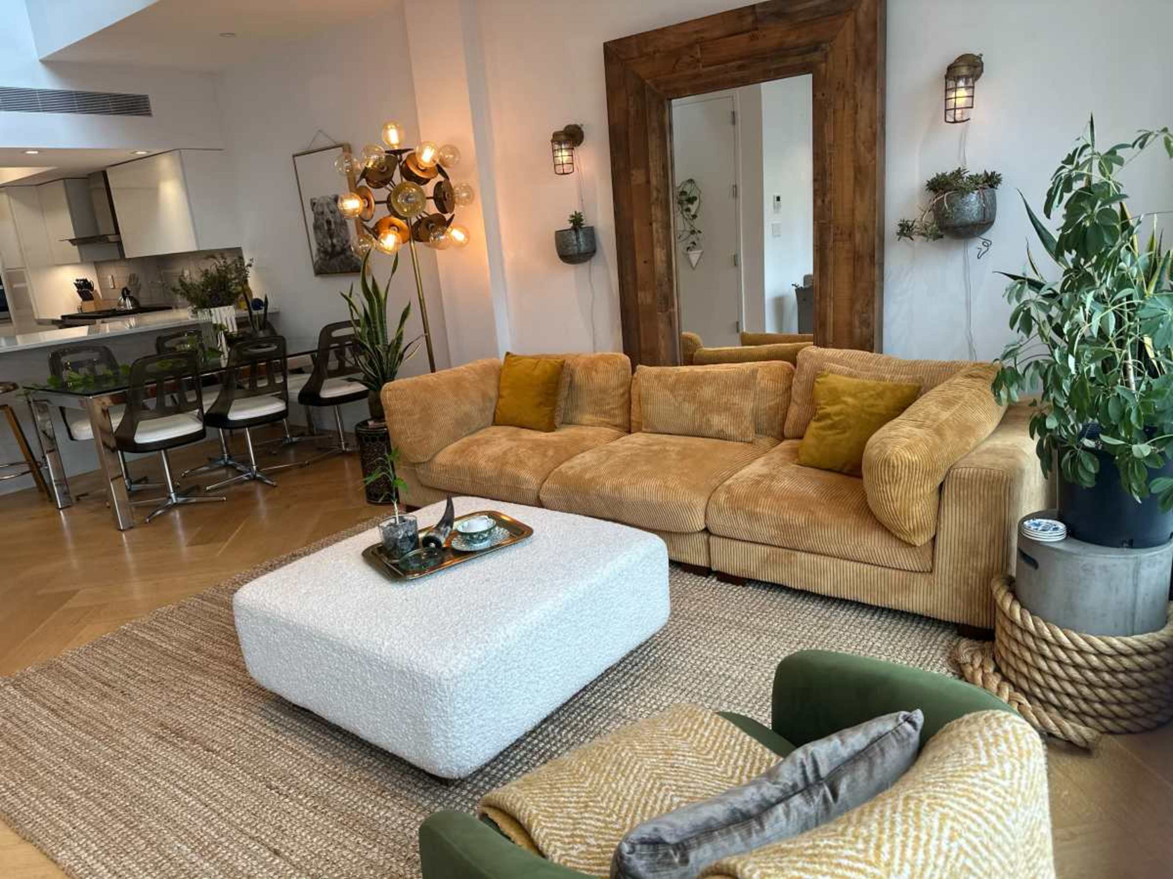 The image shows a stylish living room with a large beige sectional sofa, a white ottoman, and modern decor elements, including plants and wall art.