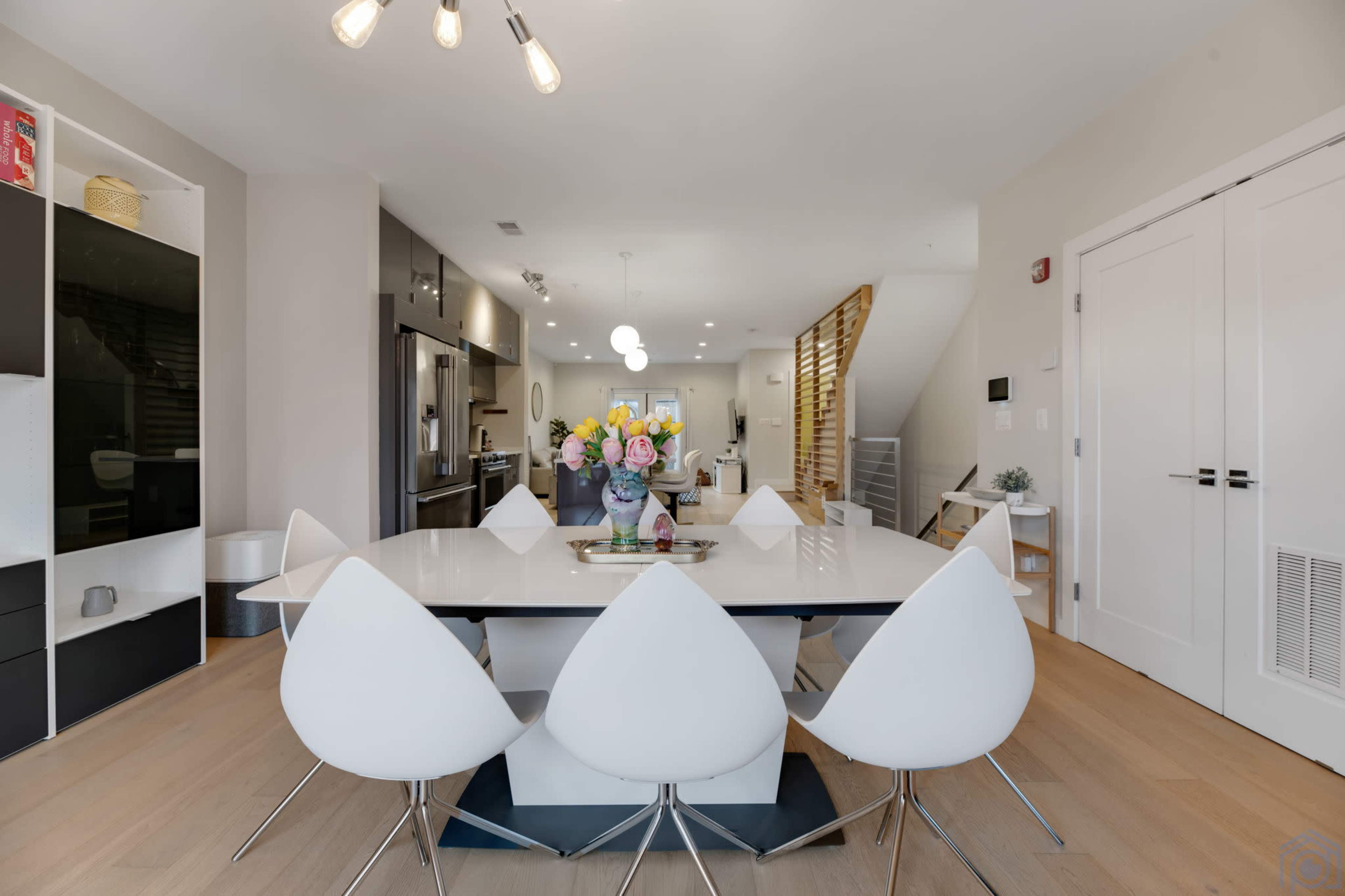 A modern dining area features a large white table surrounded by six white chairs, with a vase of flowers as the centerpiece and contemporary kitchen appliances in the background.