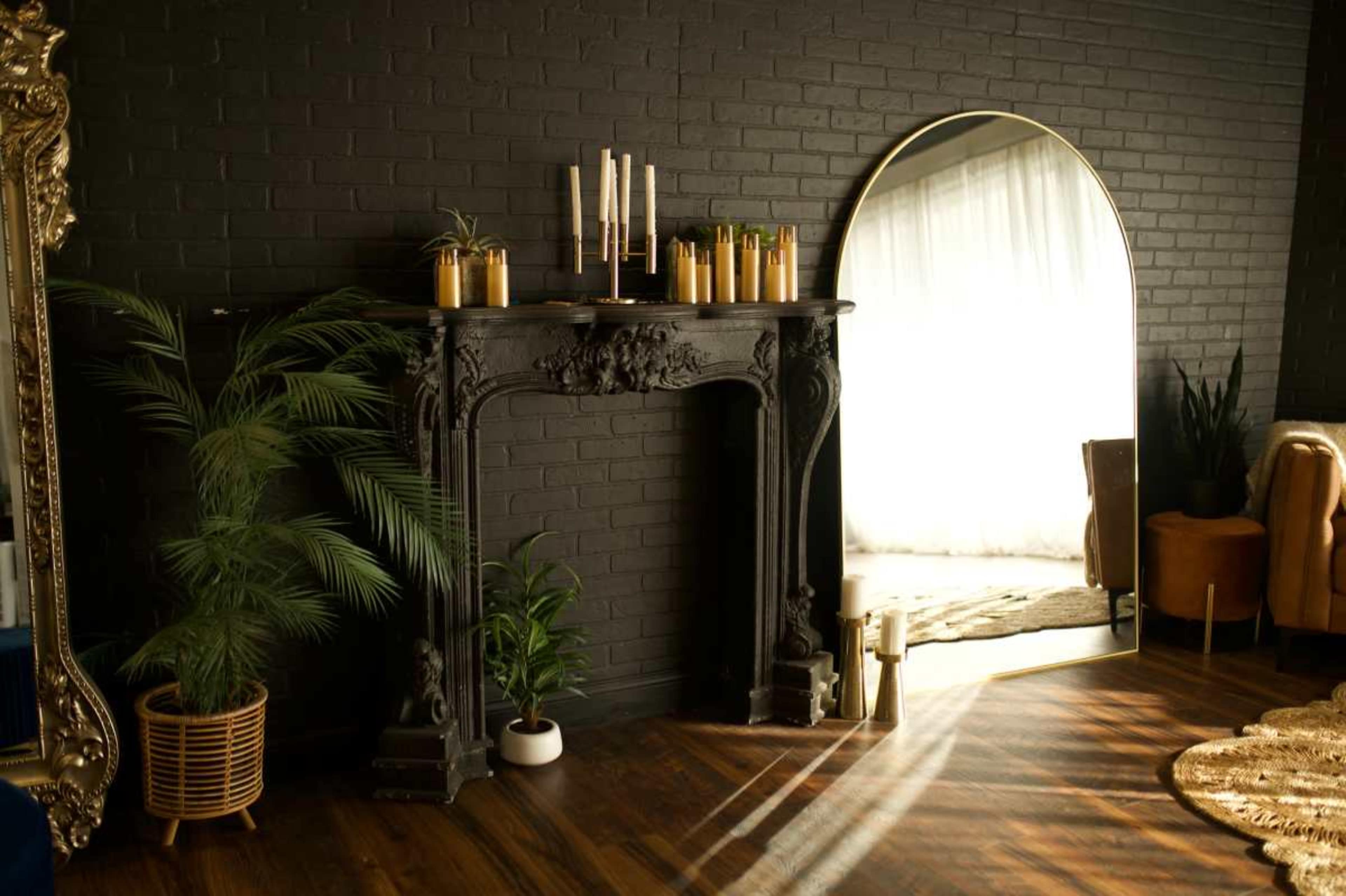 The image shows a stylish interior featuring a black-brick wall, an ornate fireplace adorned with candles, a large arched mirror, and a cozy seating area with a plant and decorative elements.