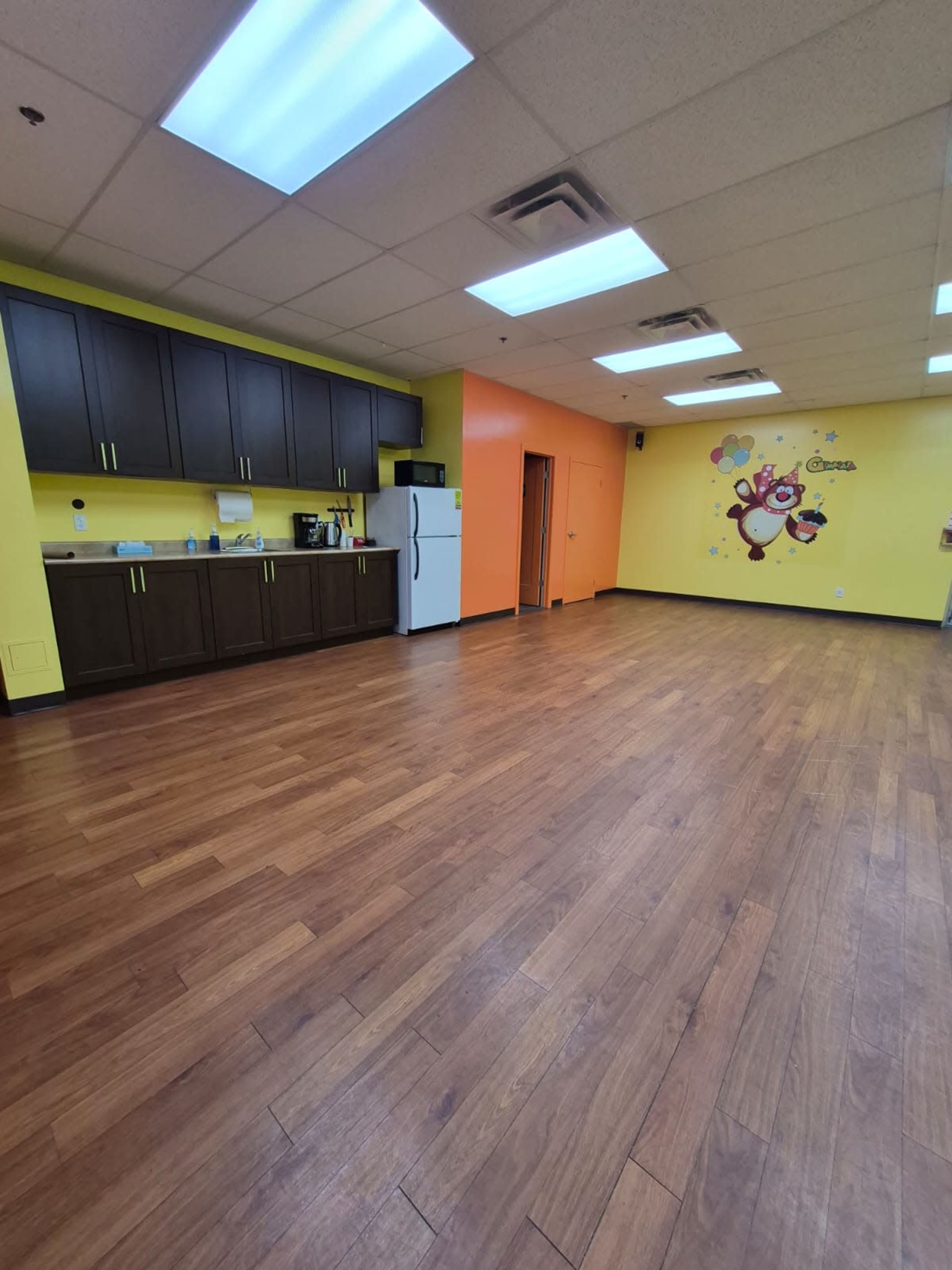 The image shows a spacious room featuring wooden flooring, yellow and orange walls, a kitchenette area with dark cabinets, and a playful wall mural.
