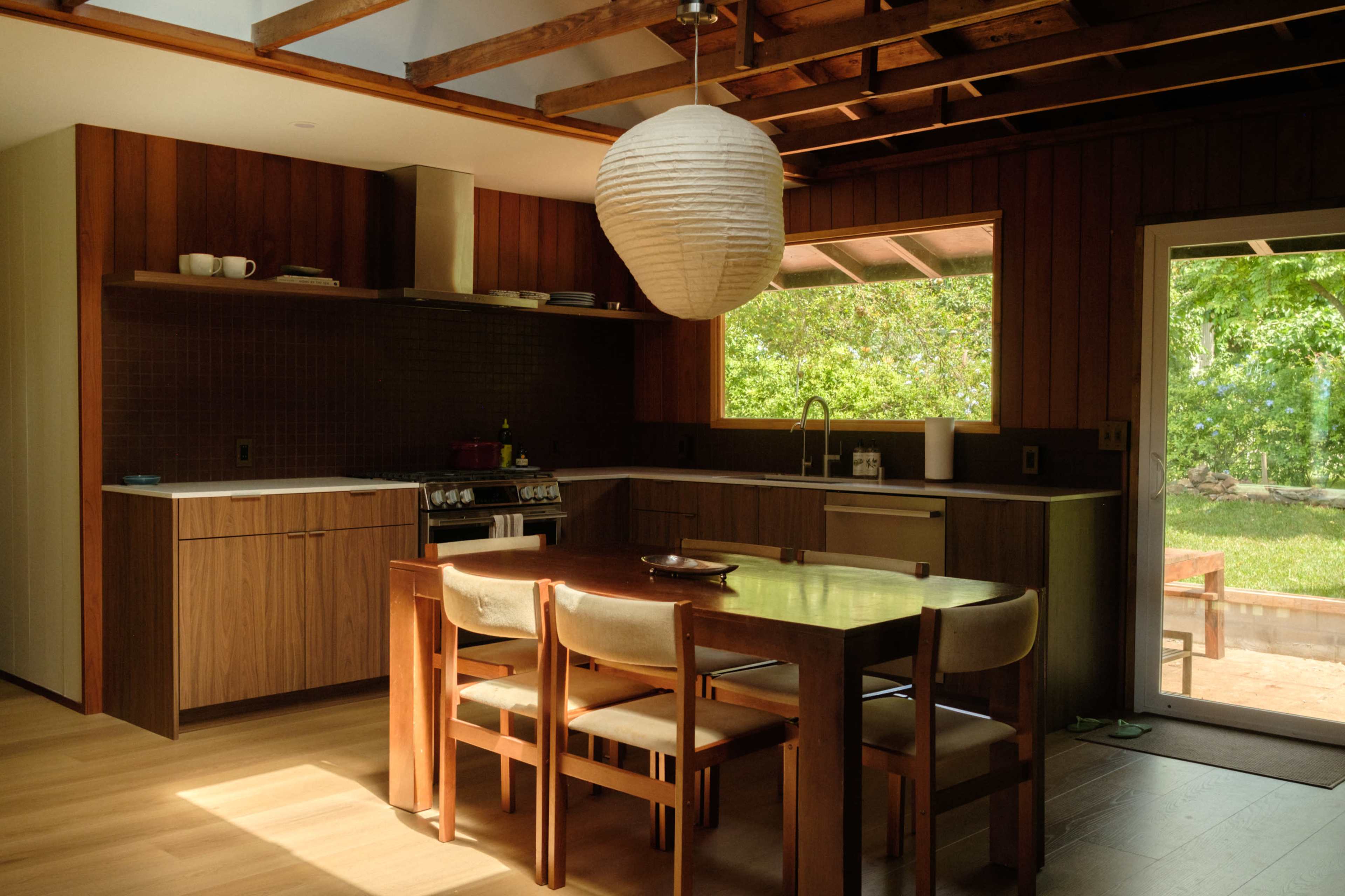 The image shows a modern kitchen and dining area with a wooden table surrounded by chairs, a light fixture, and large windows that overlook a garden.
