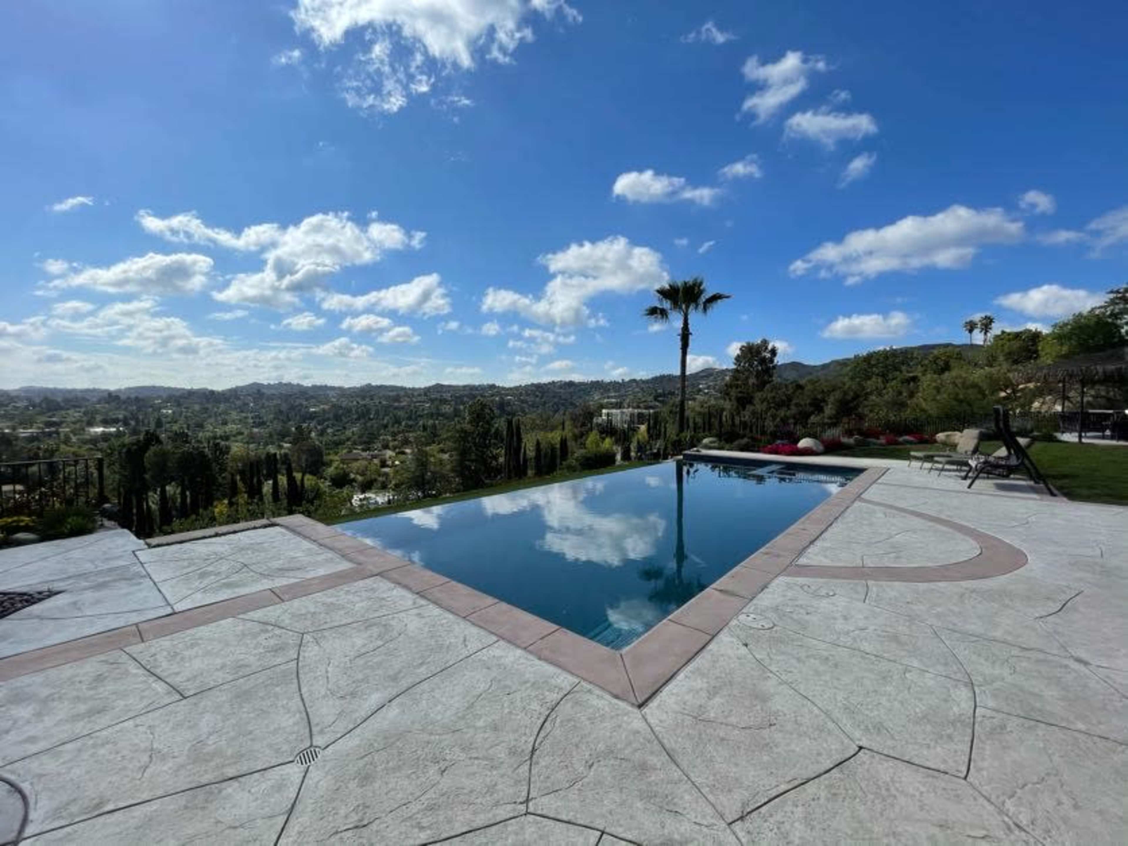 A rectangular swimming pool with a clear reflection, surrounded by a patio and overlooking a lush green landscape under a partly cloudy sky.