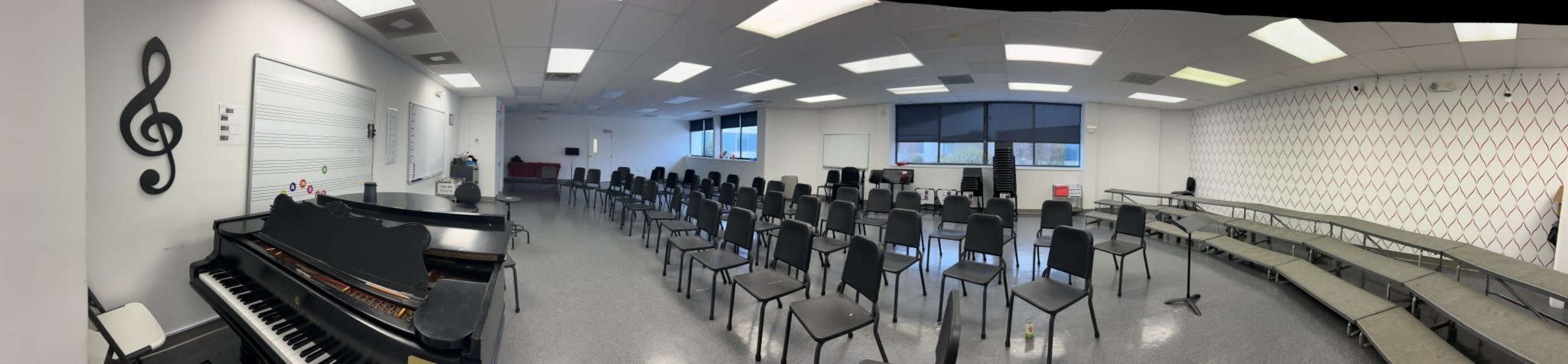 The image shows a spacious music room with a piano on the left, rows of black chairs arranged facing a large window, and a wall with musical notation symbols.