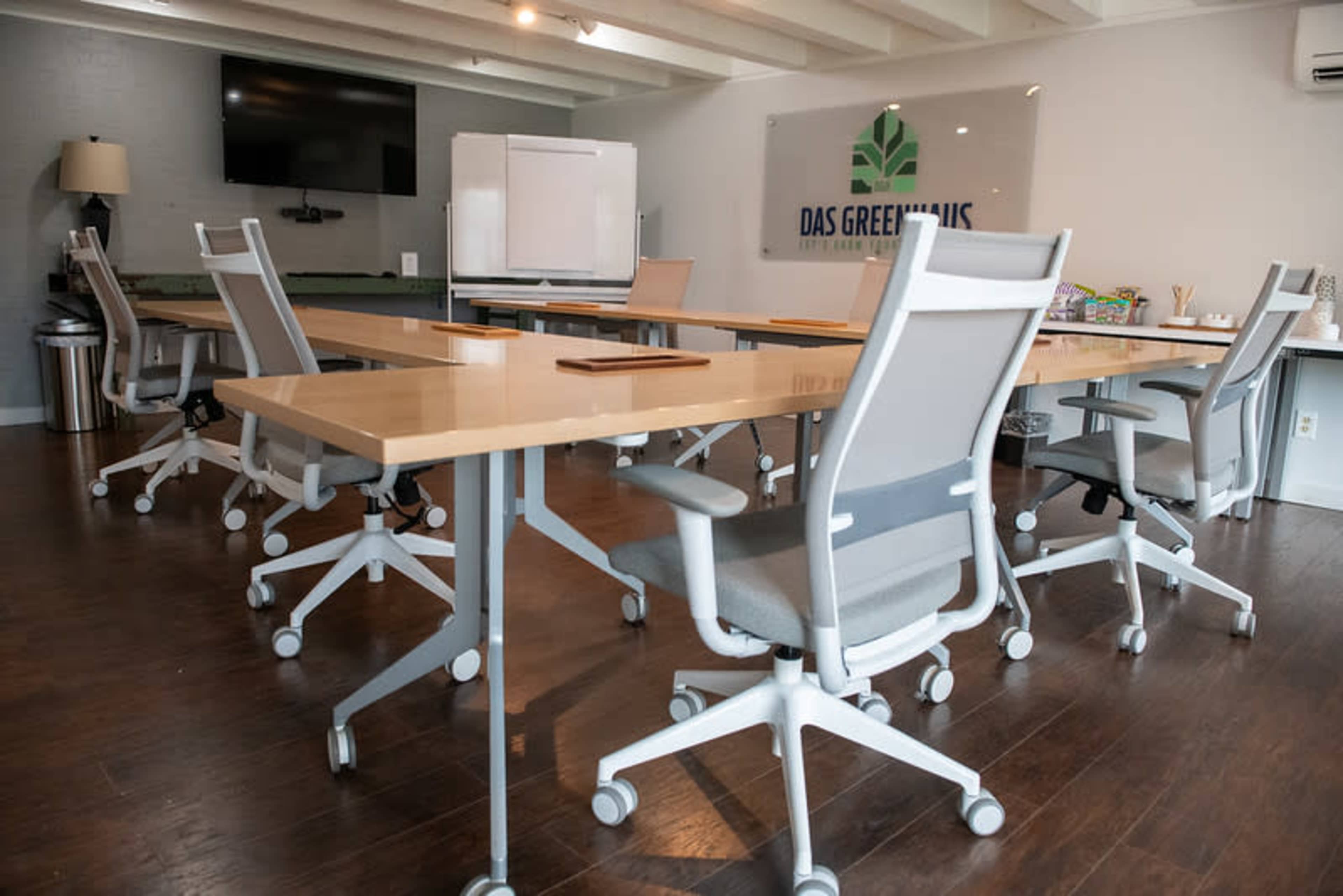 A modern conference room features rectangular tables set up with ergonomic chairs, a large screen on the wall, and a whiteboard in a well-lit environment.