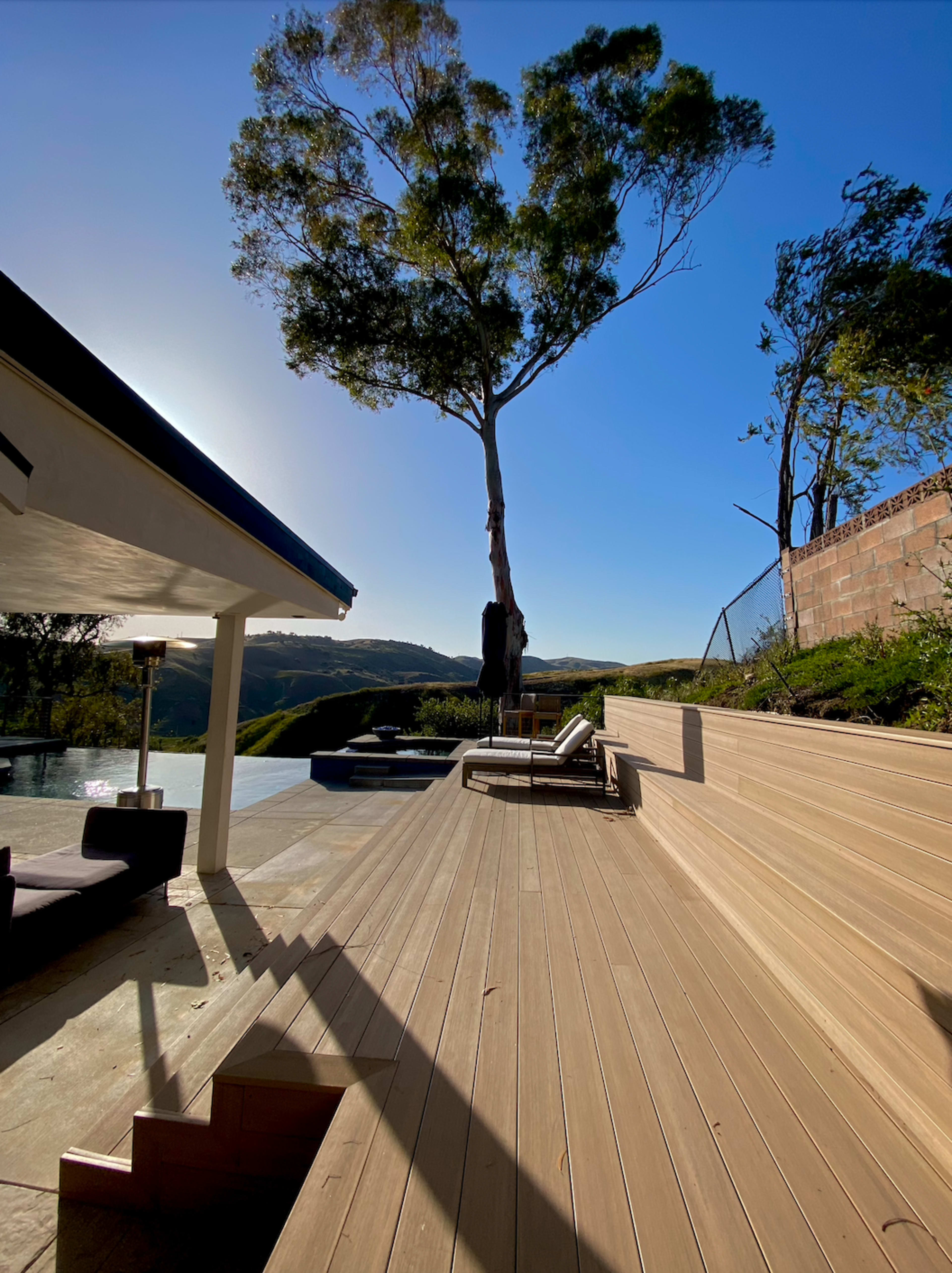 The image shows a modern outdoor deck with steps leading down to a pool area, framed by trees and hills under a clear blue sky.