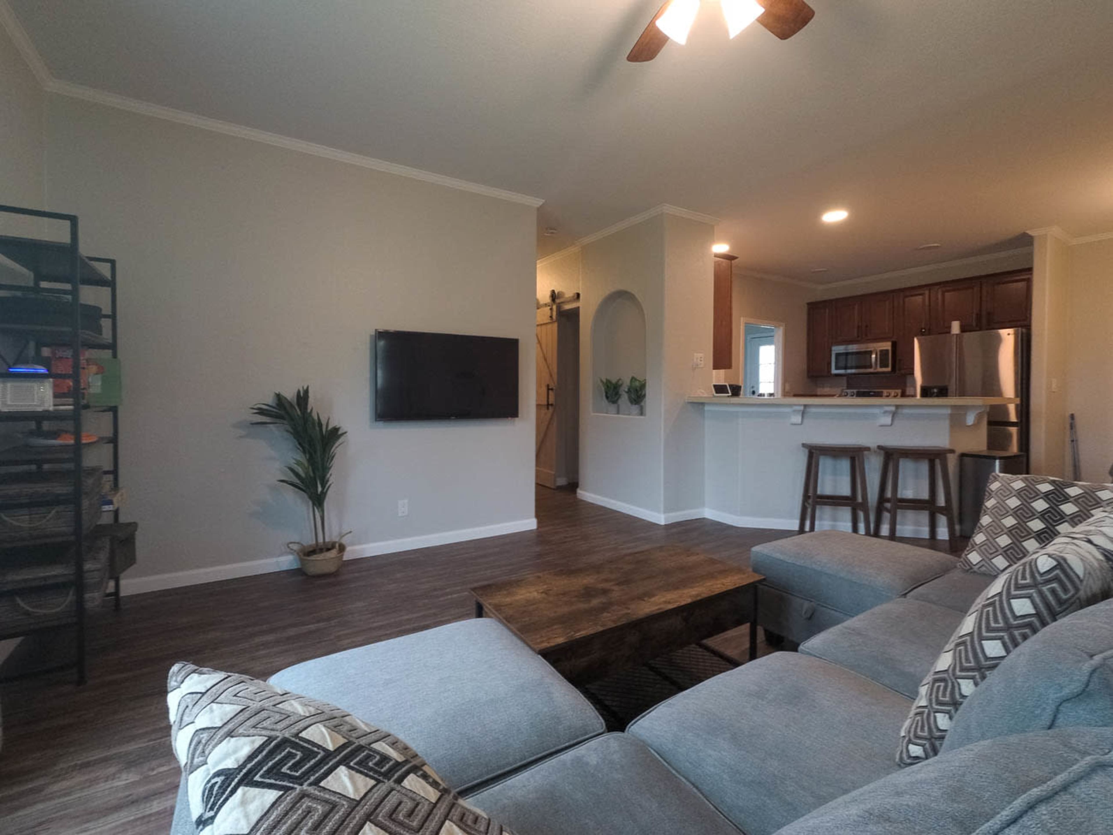 A modern living room with a sectional sofa, a wooden coffee table, and a wall-mounted TV, connected to an open kitchen area with bar stools and wooden cabinetry.