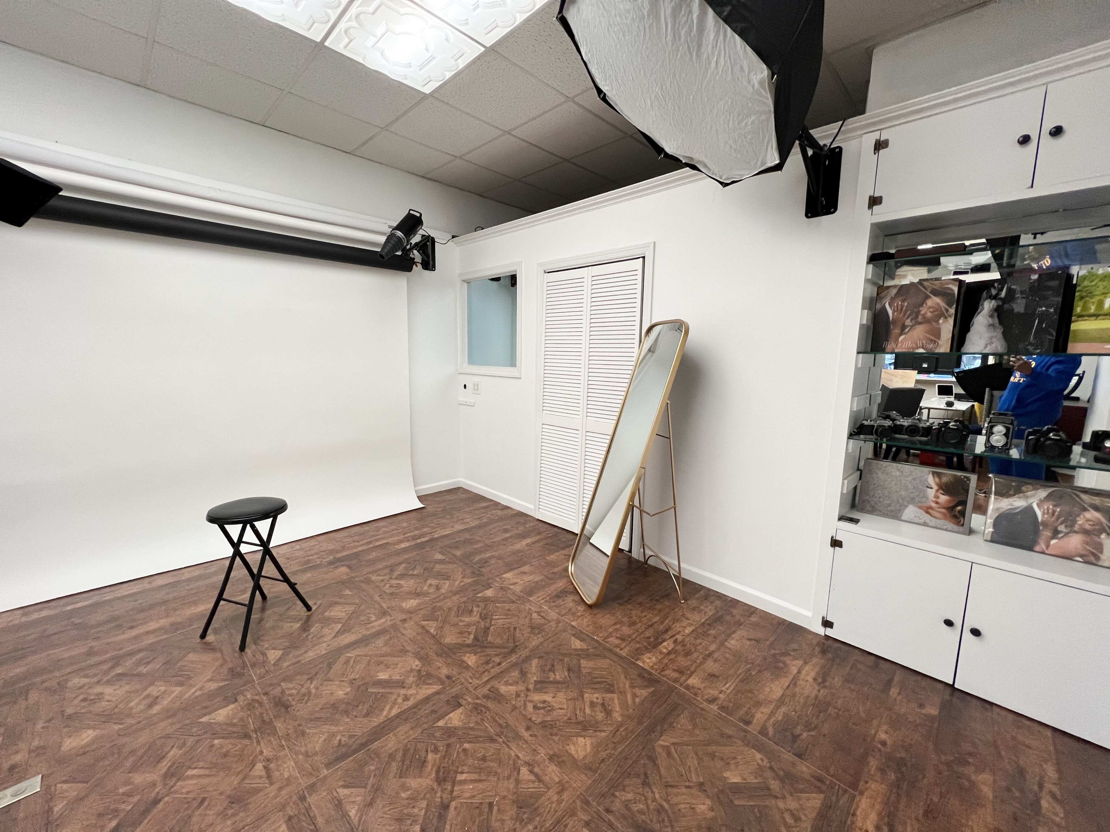 A photography studio with a blank backdrop, a stool in the center, a mirror against the wall, and storage cabinets containing images.