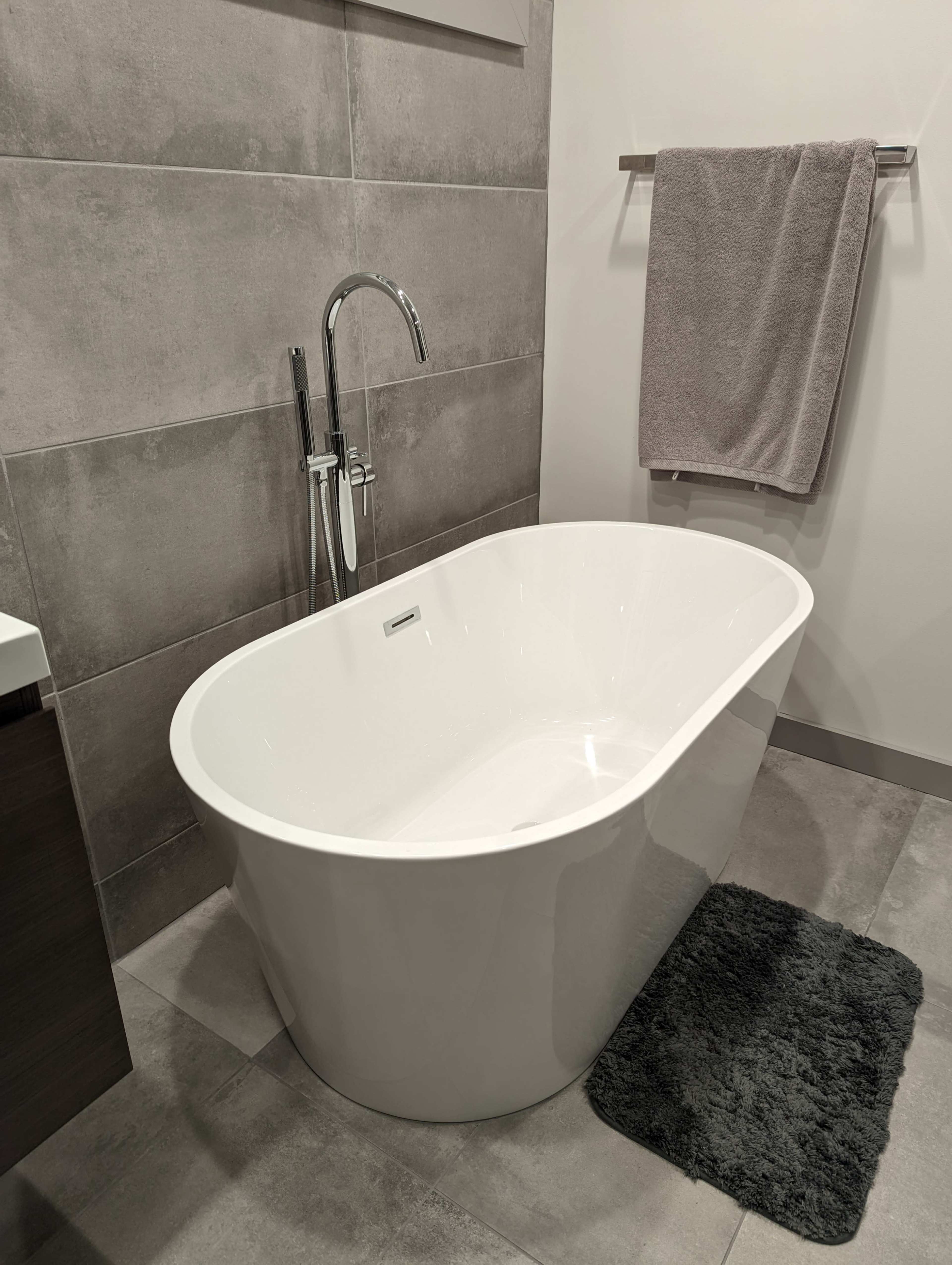 A modern bathroom featuring a freestanding white bathtub next to a towel hanging on a rack and a dark gray rug on the floor.