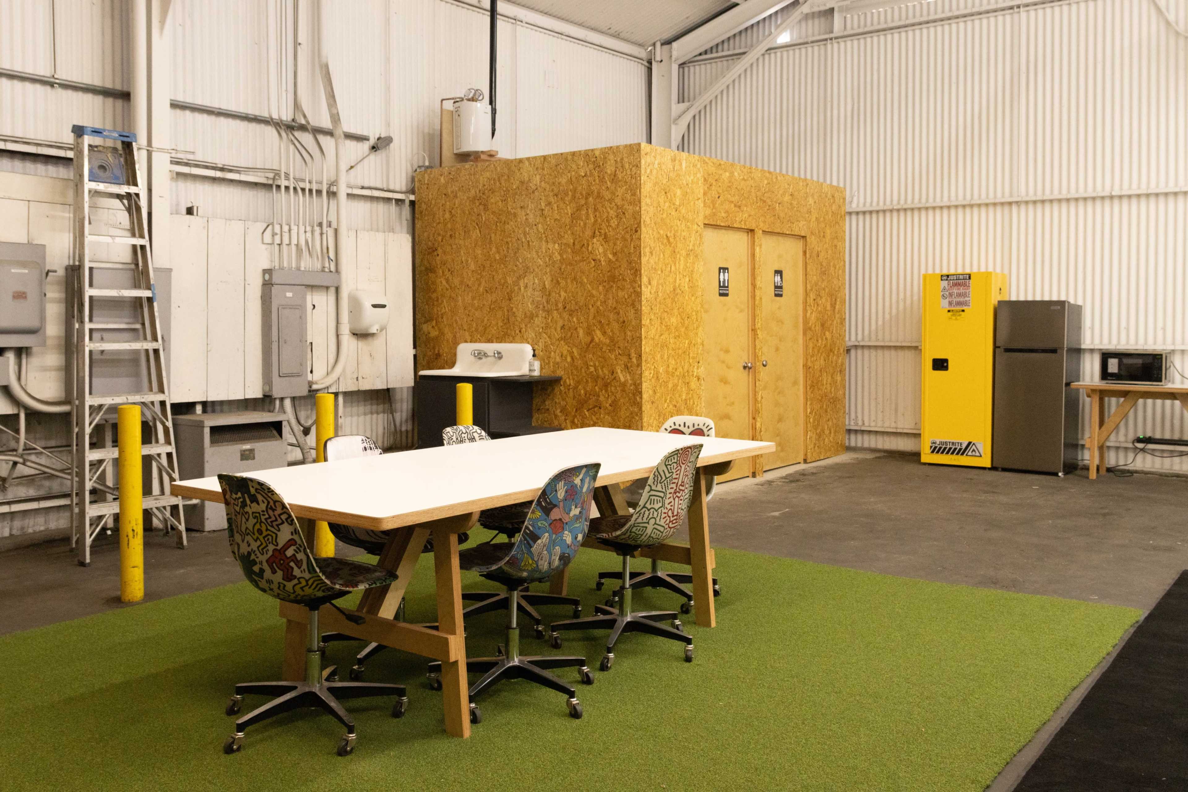 The image shows a spacious indoor area with a long table surrounded by colorful chairs, a kitchenette area, and two doors at the back, all set on a green carpet-like flooring.