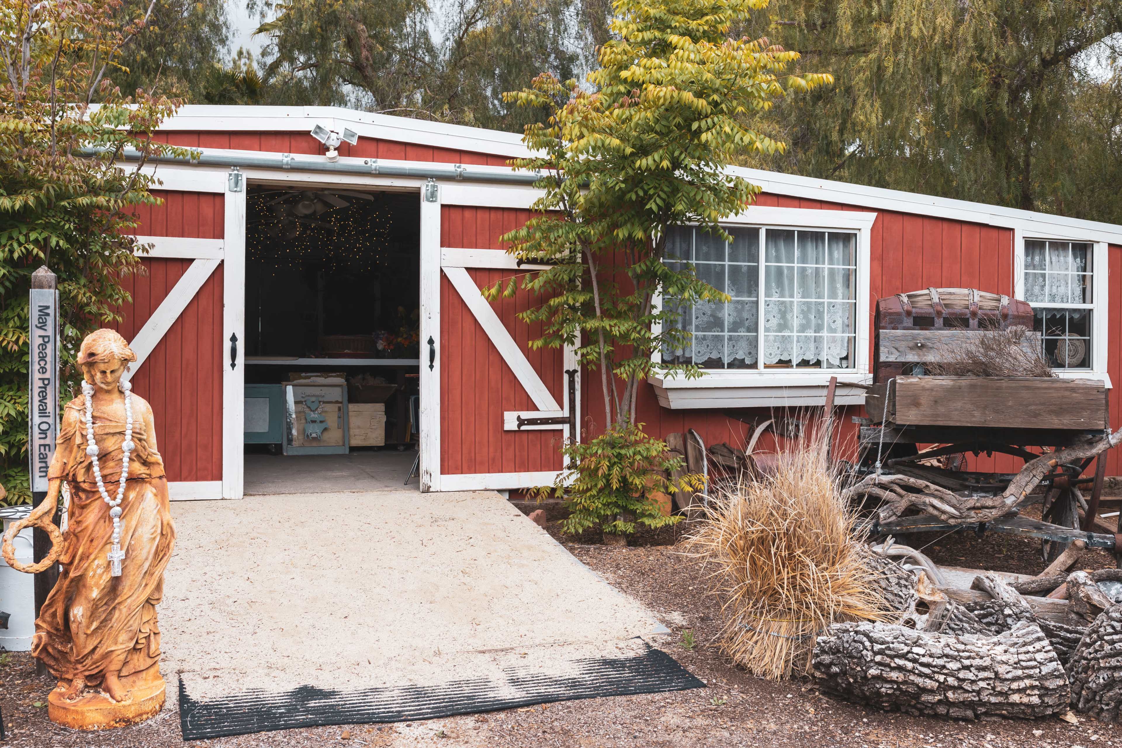 A red barn-style building with white trim features double doors, surrounded by greenery, and a statue of a figure stands to the left.