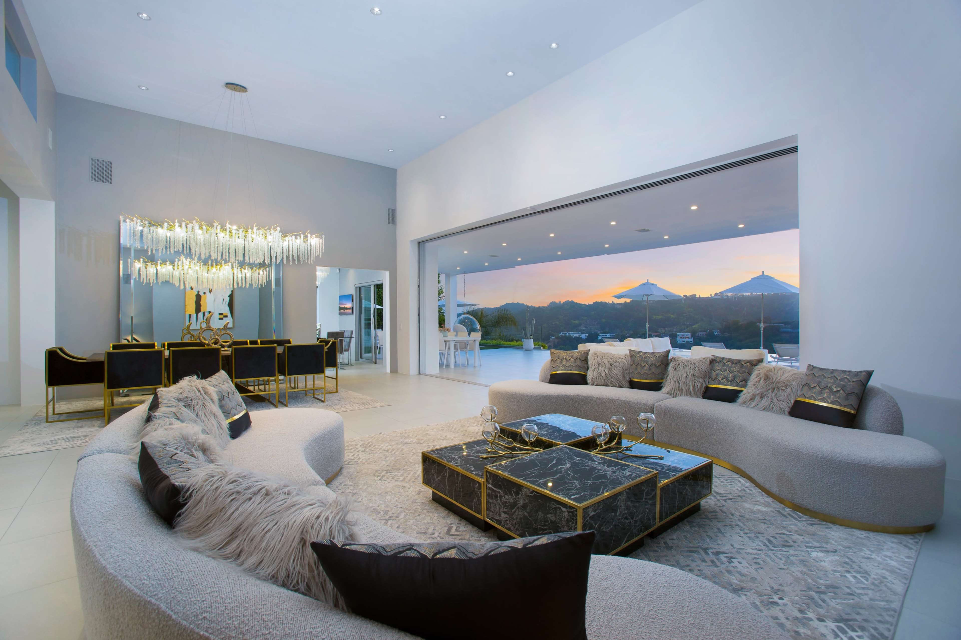 The image shows a modern living room with a curved sofa, a marble coffee table, and large glass doors leading to an outdoor view at sunset.