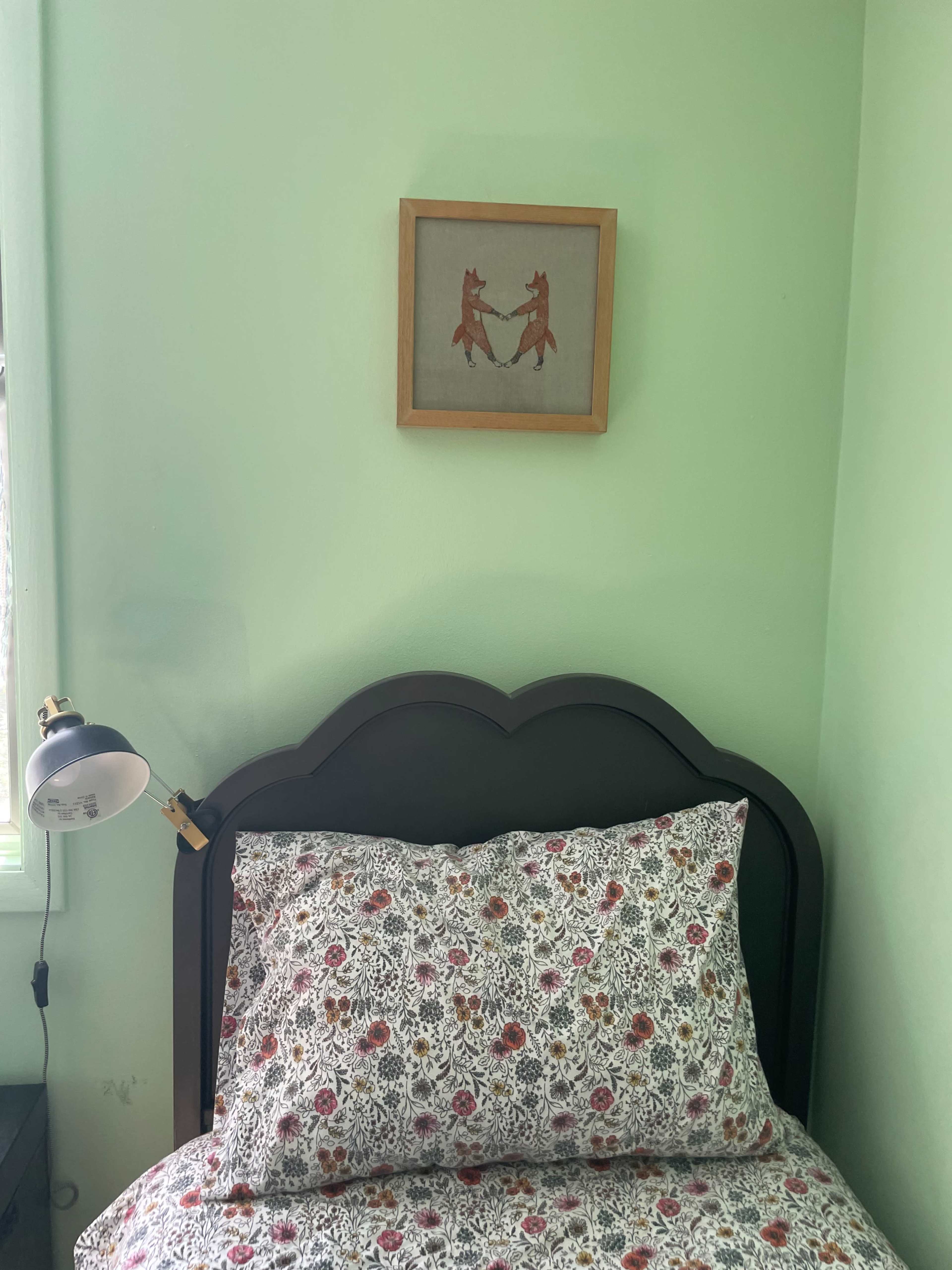 A bed with a dark headboard and floral-patterned pillows is positioned against a light green wall, featuring a framed artwork above it and a lamp on the bedside table.