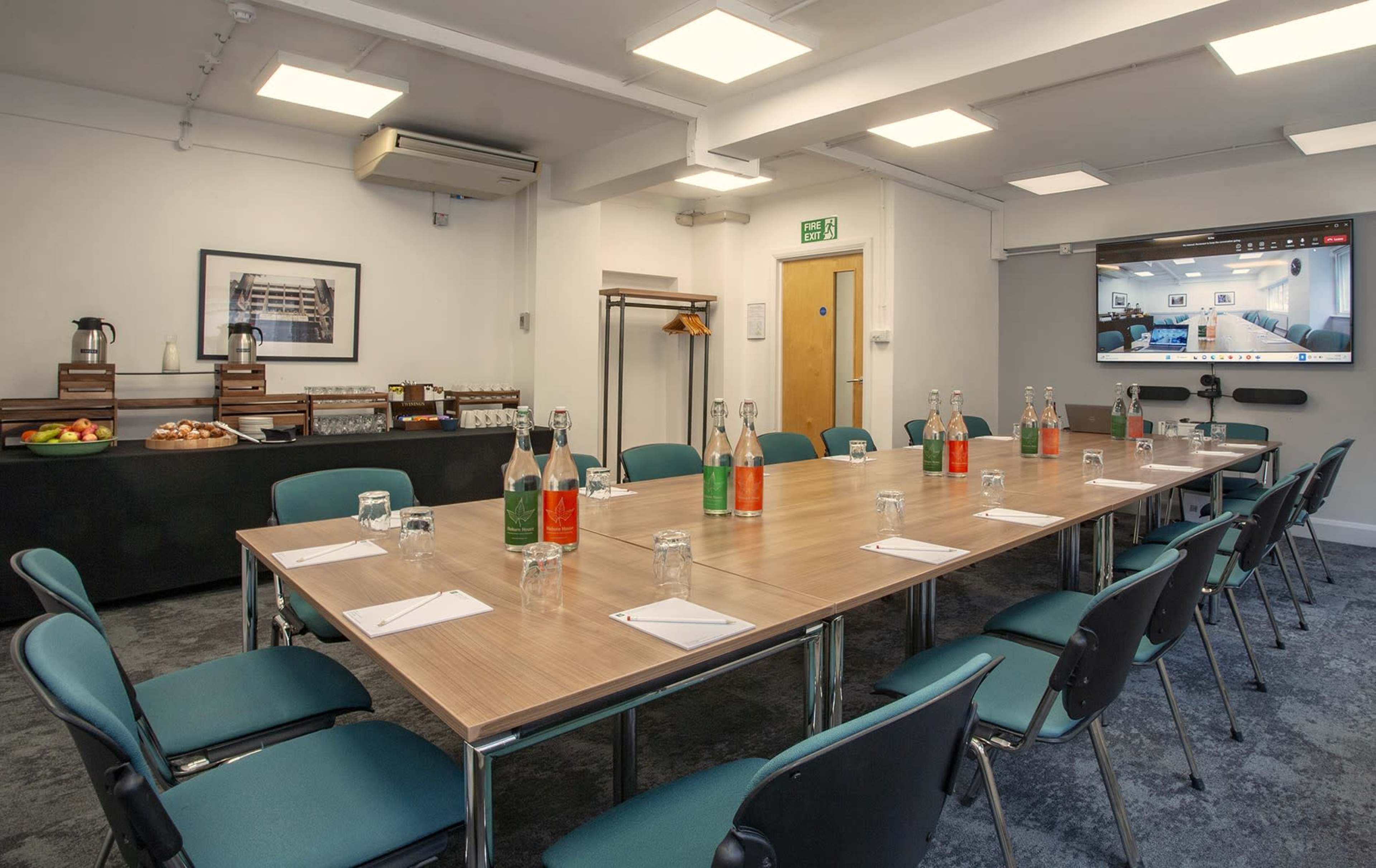 A meeting room is set with a large table surrounded by green chairs, featuring water bottles, notepads, and a screen displaying virtual meeting participants.