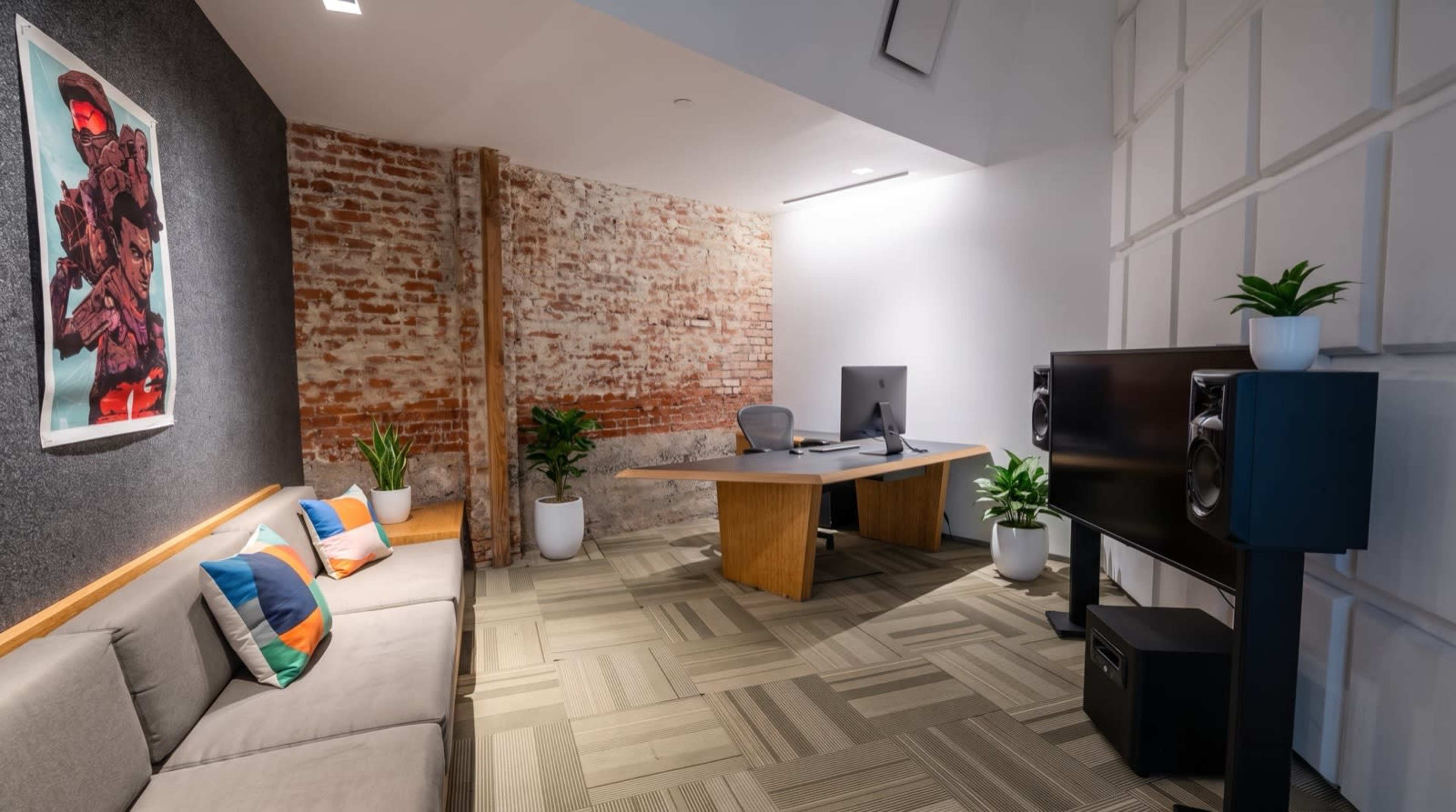 The image shows a modern office space featuring exposed brick walls, a large wooden desk, a computer setup, a gray sofa, and potted plants.