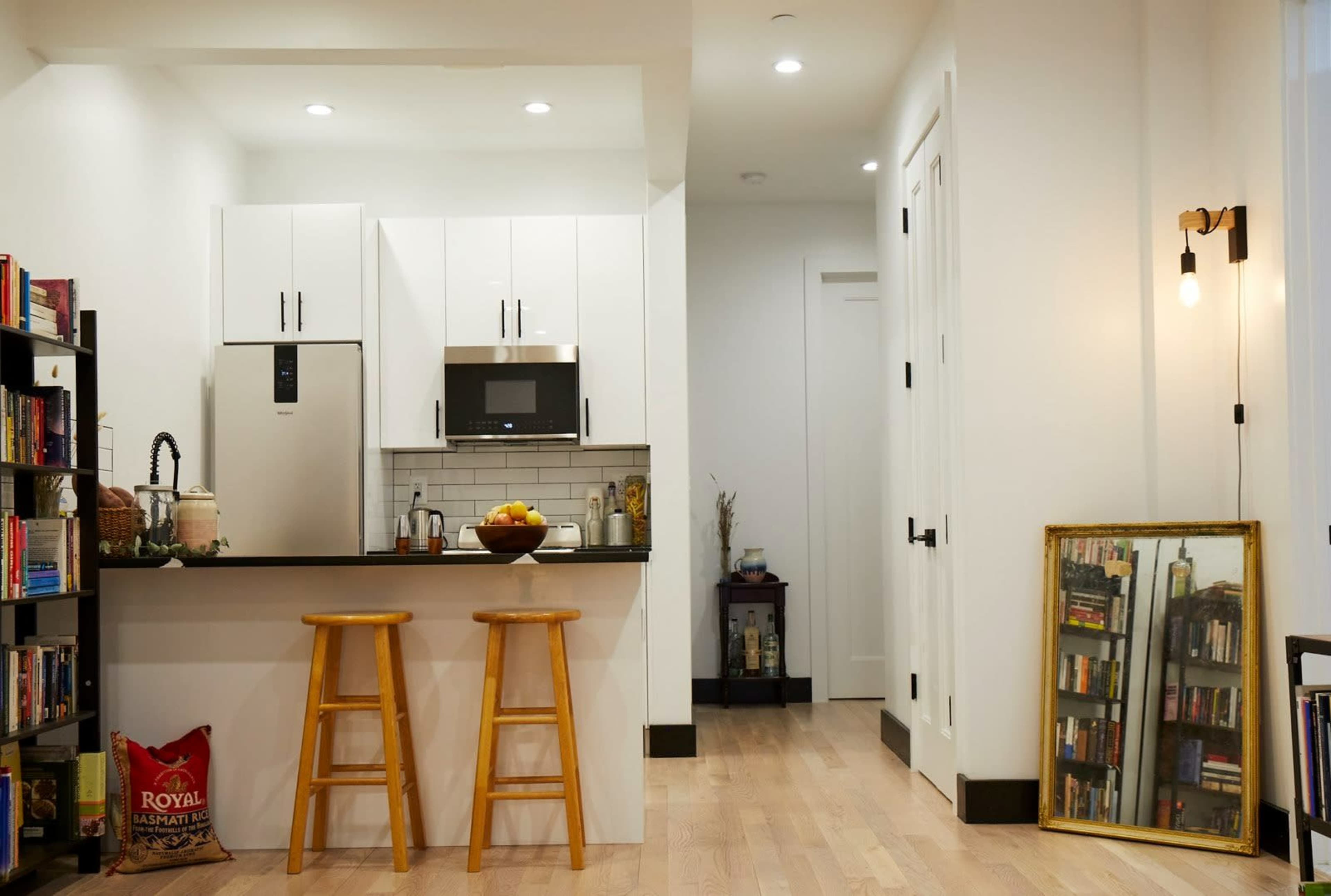 A modern kitchen with white cabinets, a small dining area featuring two wooden stools, and a reflecting mirror next to a bookshelf.