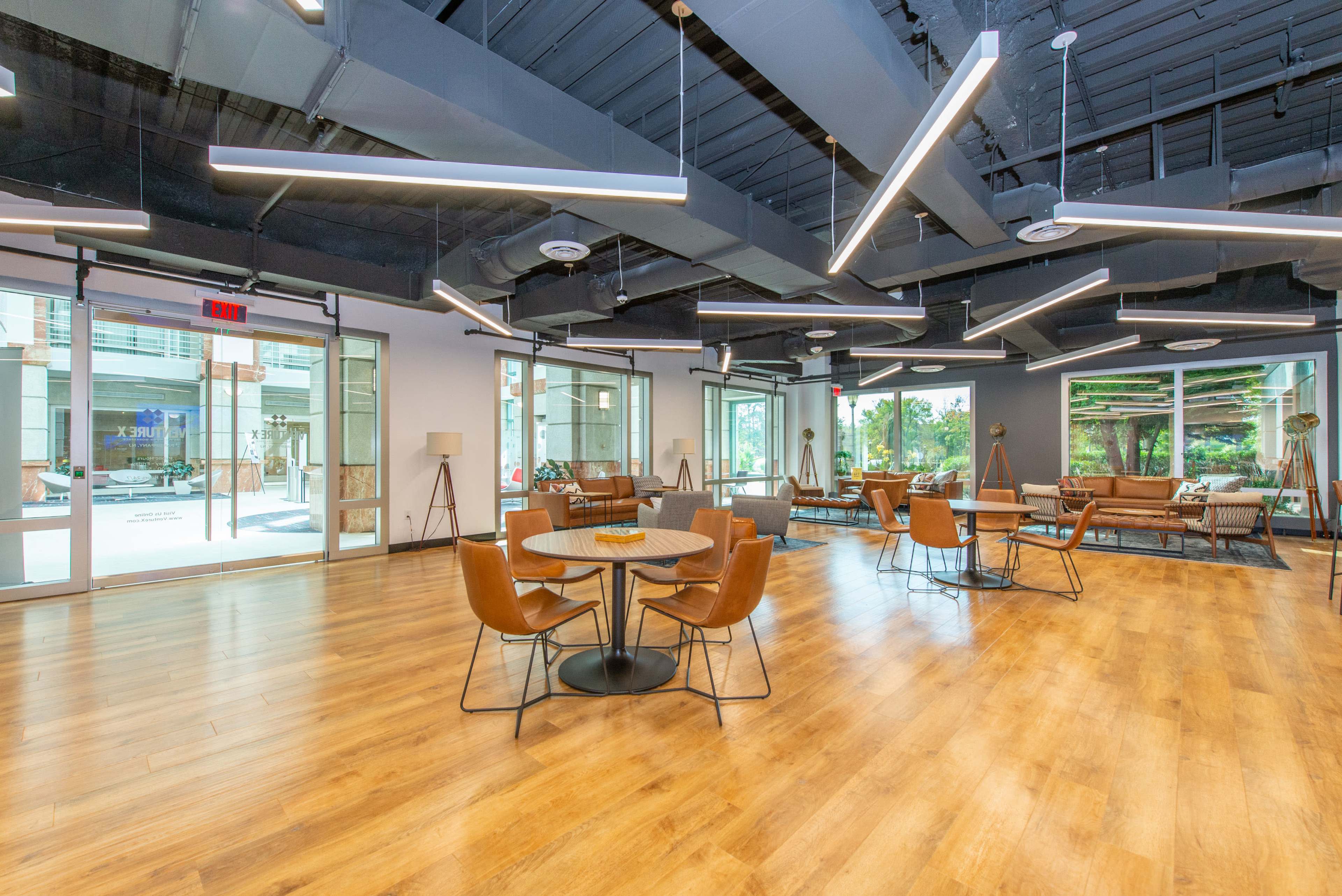 The image shows a modern indoor space with wooden flooring, several seating areas, and large windows that open to an outdoor area.