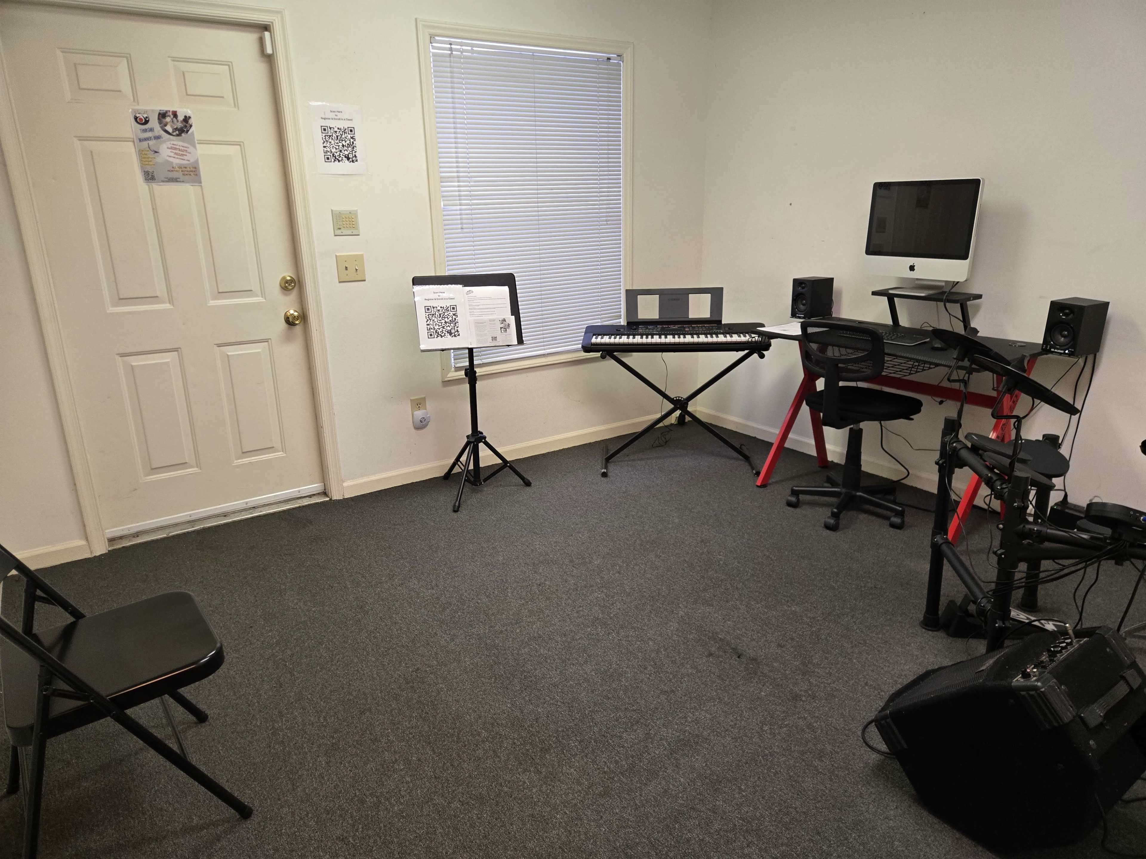 The image shows a small room with a keyboard, computer setup, music stands, and a few chairs arranged around the space.