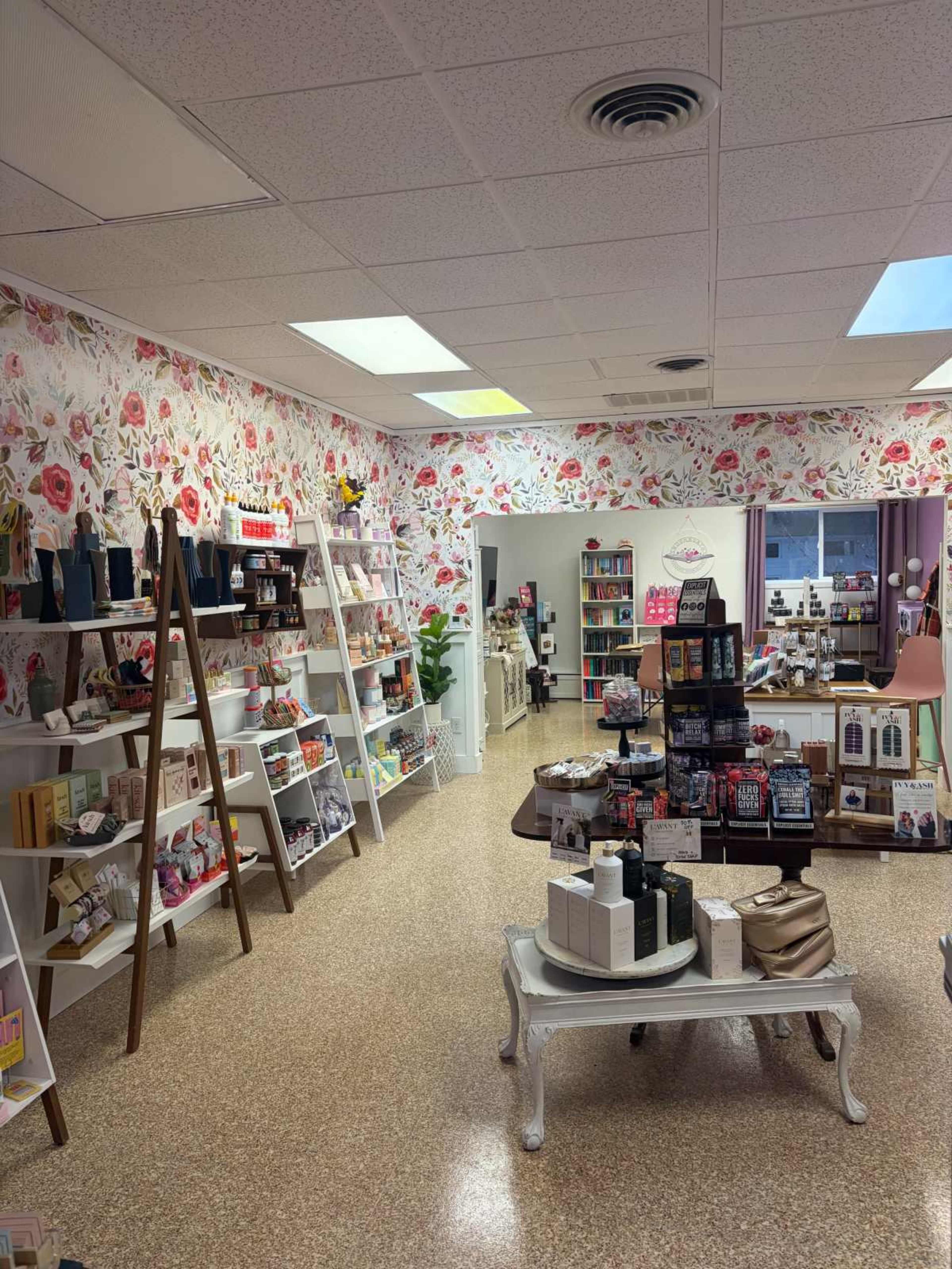 The image shows the interior of a retail store featuring a floral wallpaper, various shelves stocked with beauty and personal care products, and a seating area with a table in the center.