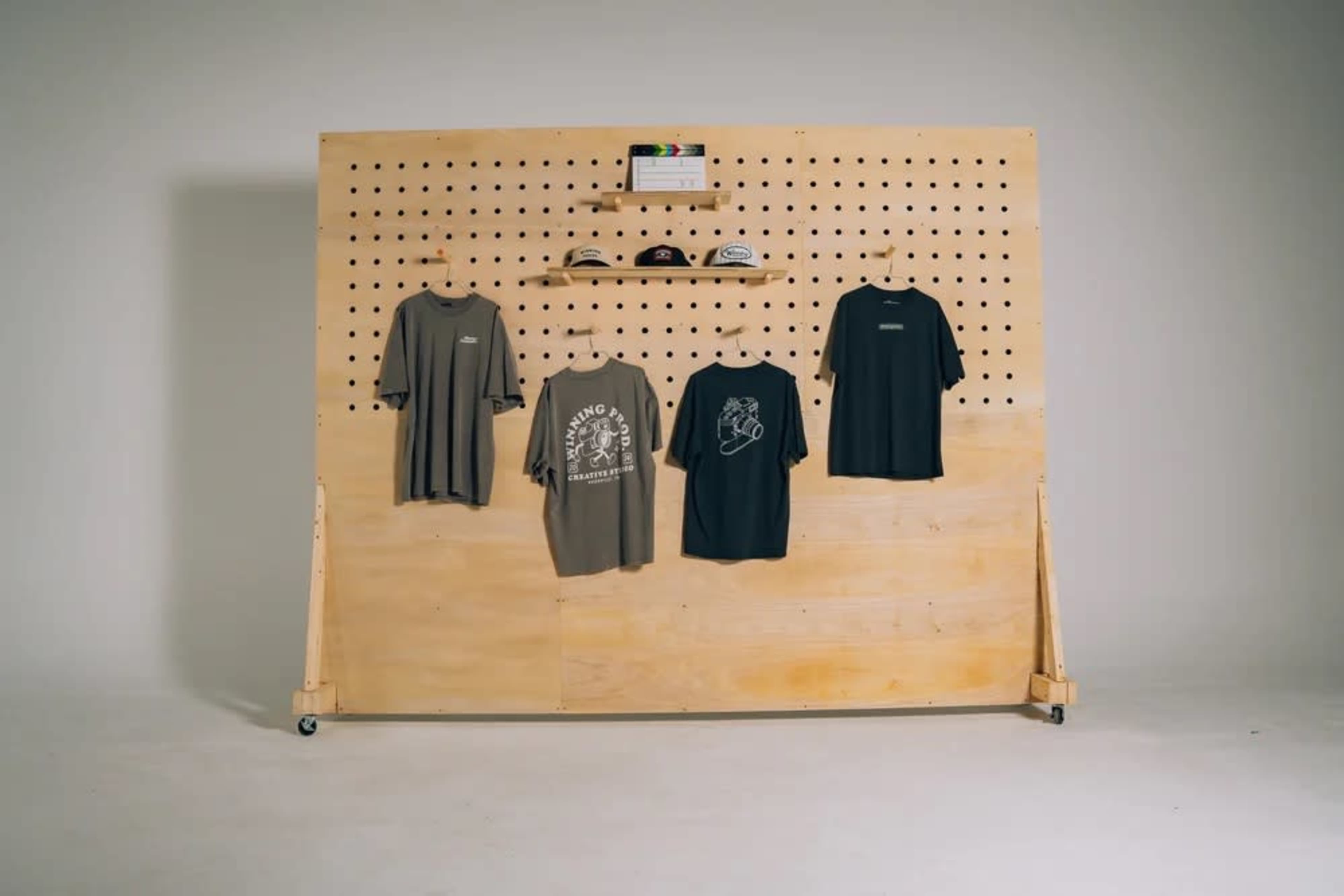 The image shows a display featuring five t-shirts hanging on a wooden pegboard wall, with some caps resting on a shelf above them.