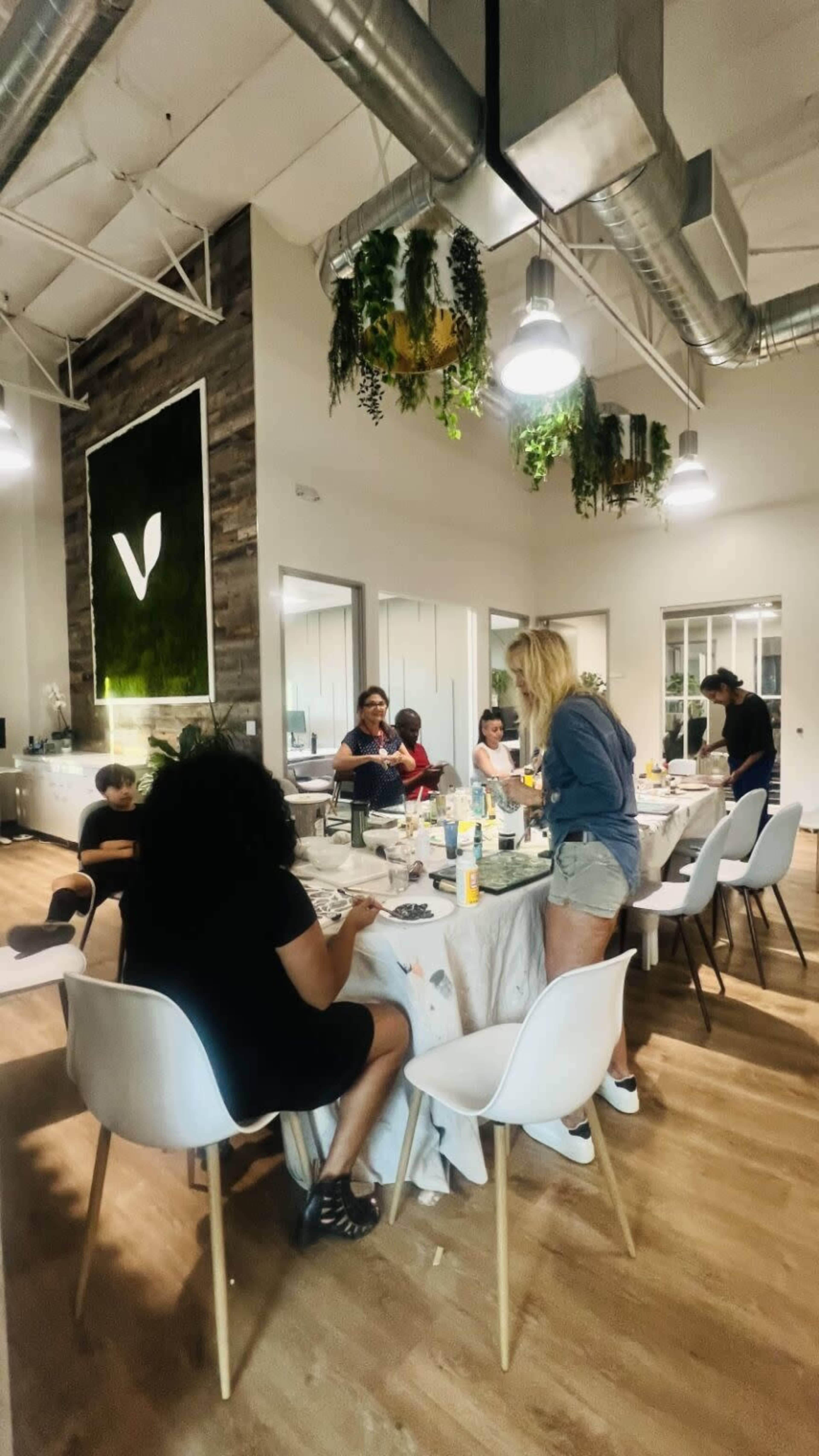 A group of people are engaged in conversation around a dining table in a modern office space with greenery decor.