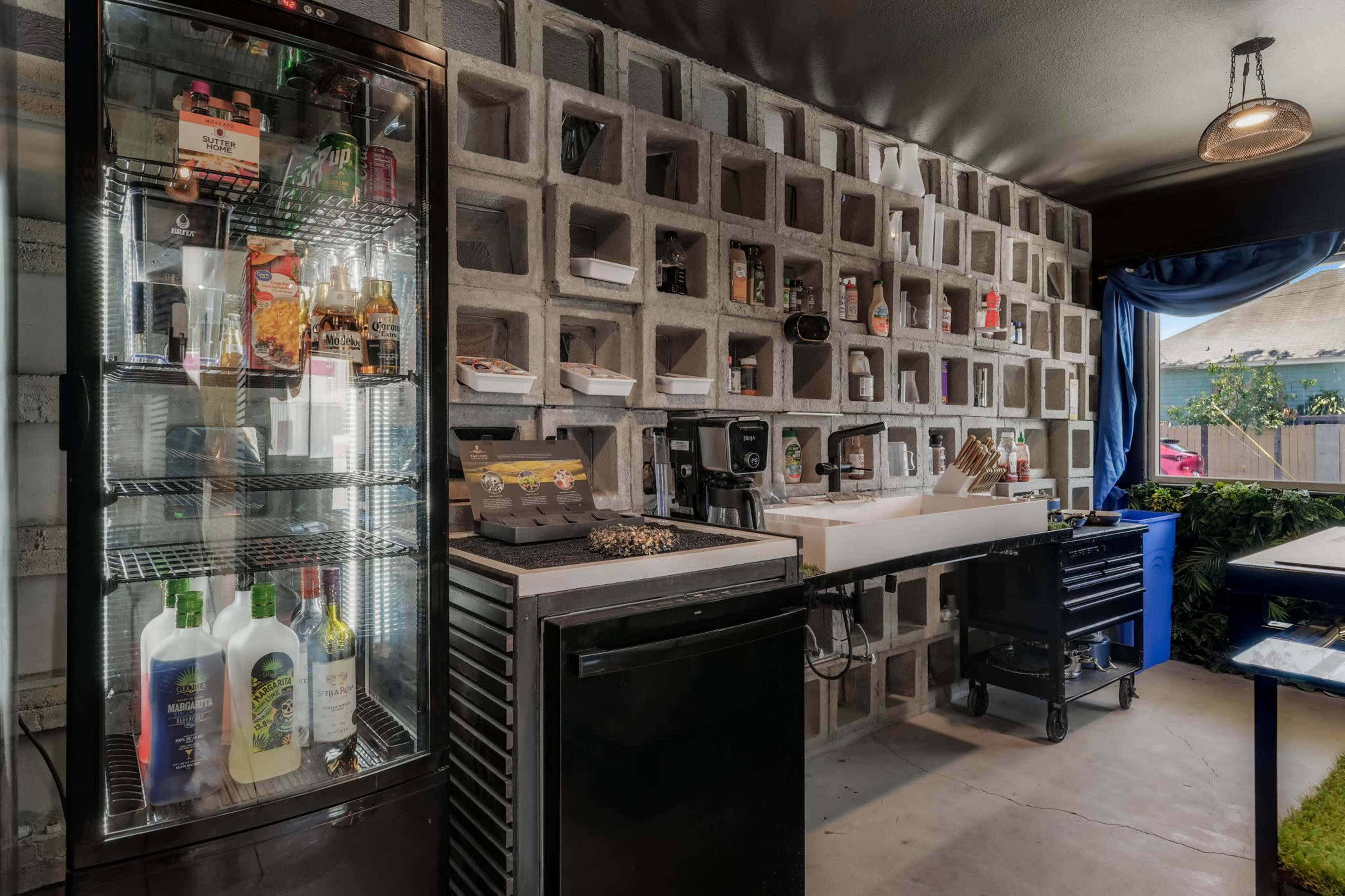 The image shows a modern bar setup featuring a glass-fronted refrigerator, a countertop with coffee-making equipment, and a wall made of decorative concrete blocks displaying various bottles and supplies.