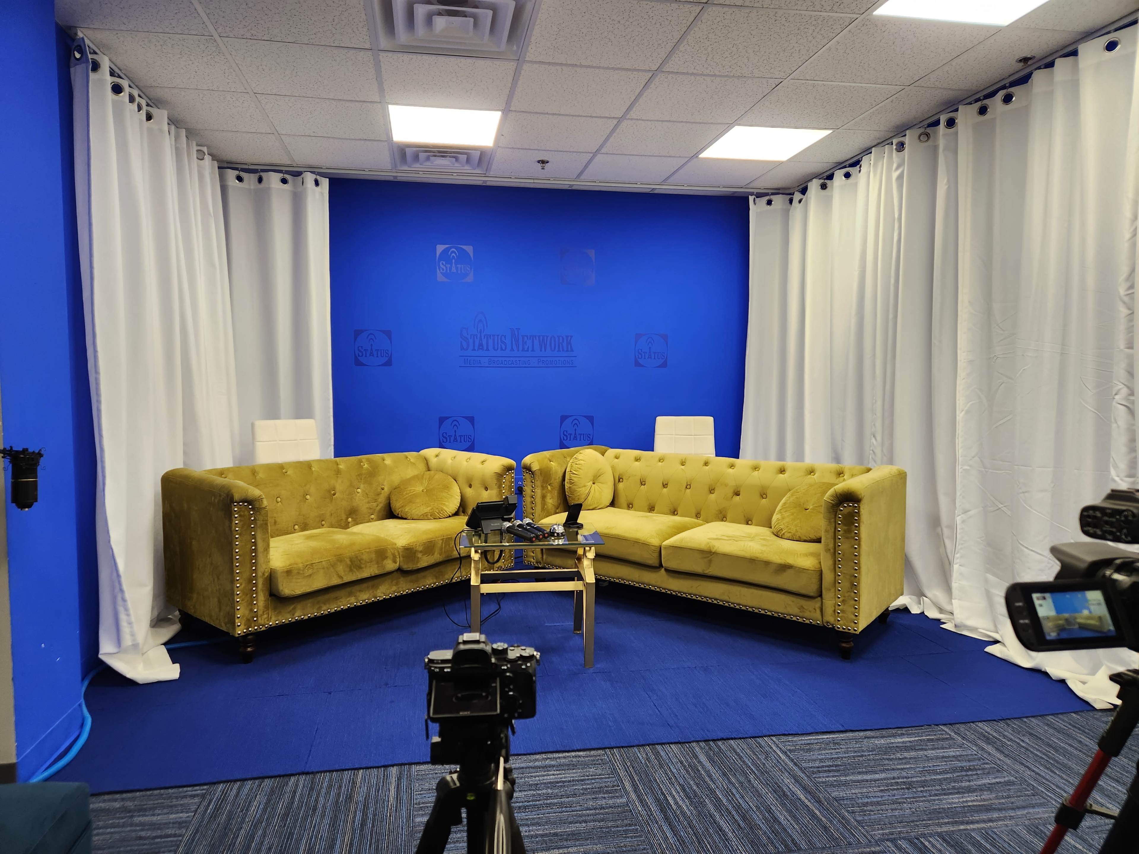 A television studio setup with two yellow tufted sofas facing each other on a blue carpet, surrounded by white curtains and a blue backdrop.