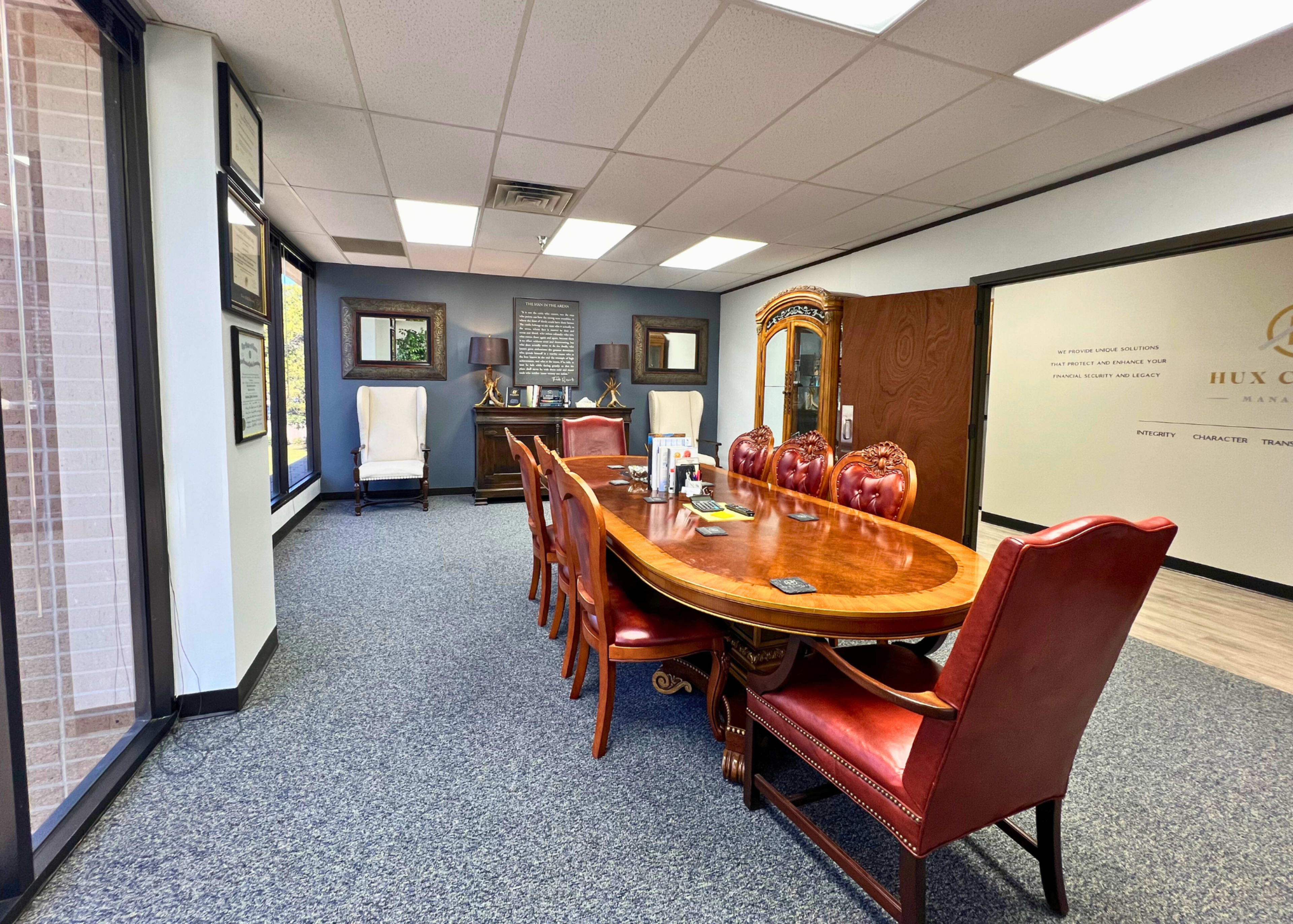 A conference room features a large wooden table surrounded by red leather chairs, with framed artwork and a cabinet visible in the background.