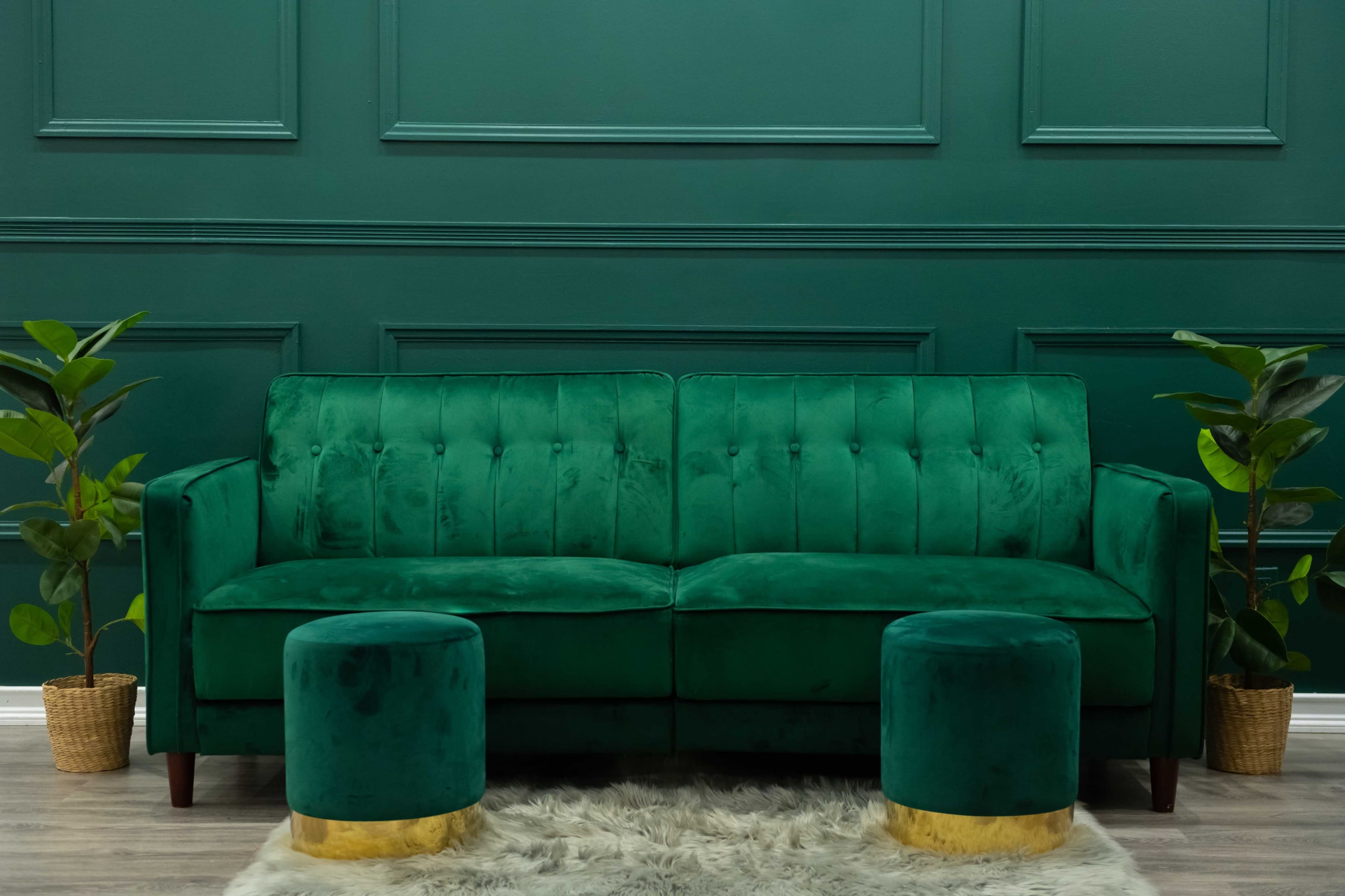 A green velvet sofa with tufted detailing is placed against a dark green wall, flanked by two potted plants and two cylindrical stools.