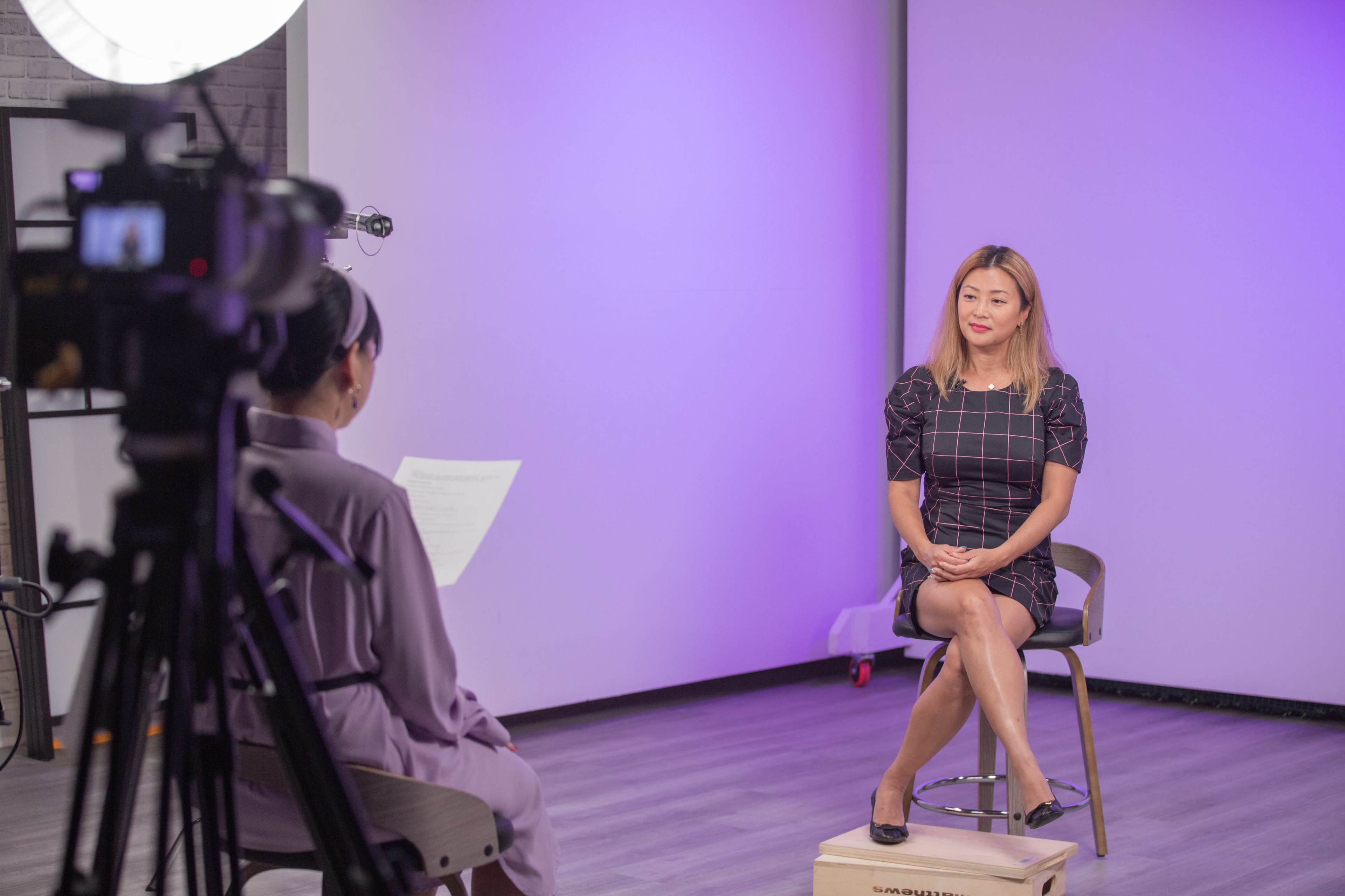 A woman sits in a chair, facing a camera setup, while another person reads from a script.