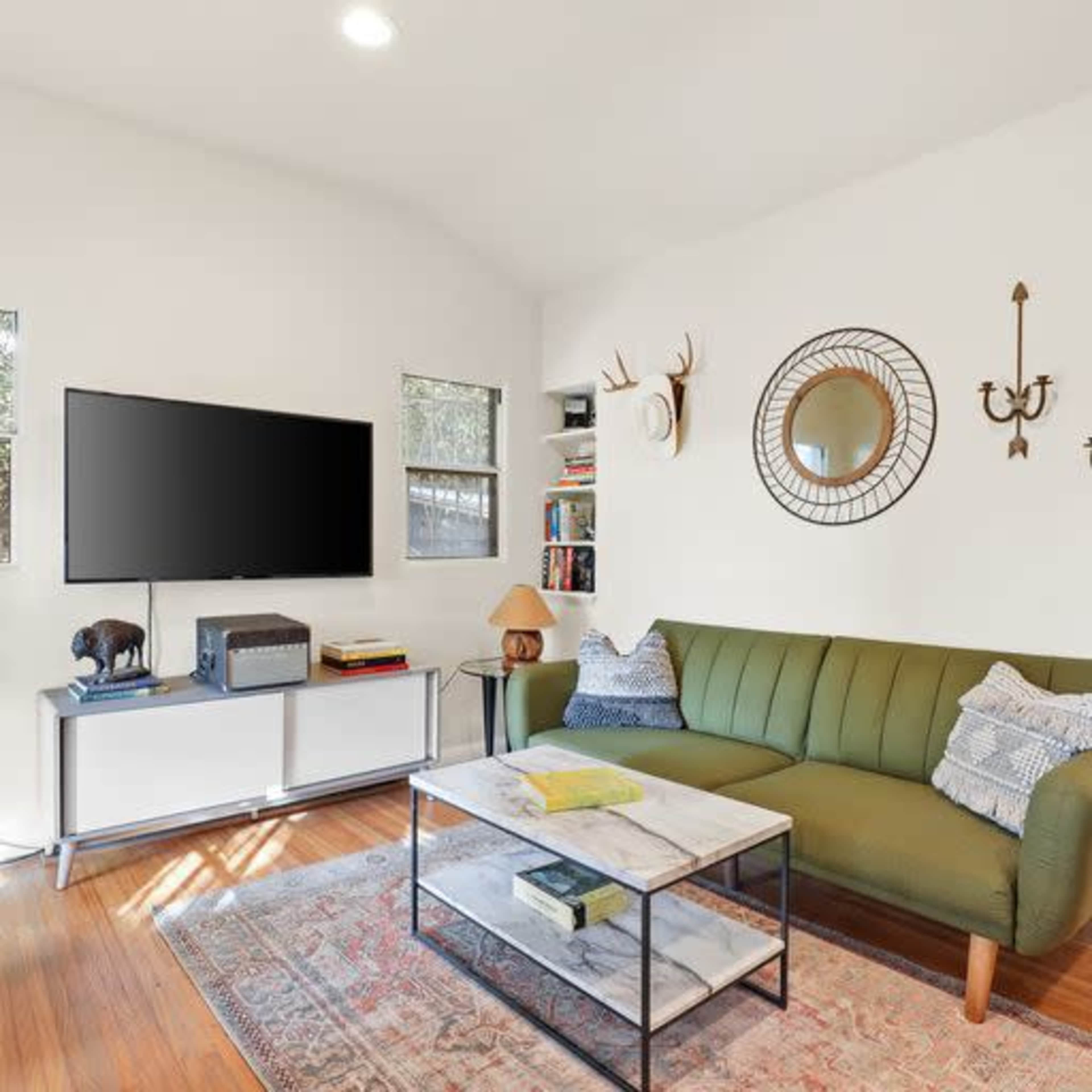 A living room with a green sofa, a coffee table, a television mounted on the wall, and shelves containing books and decor items.