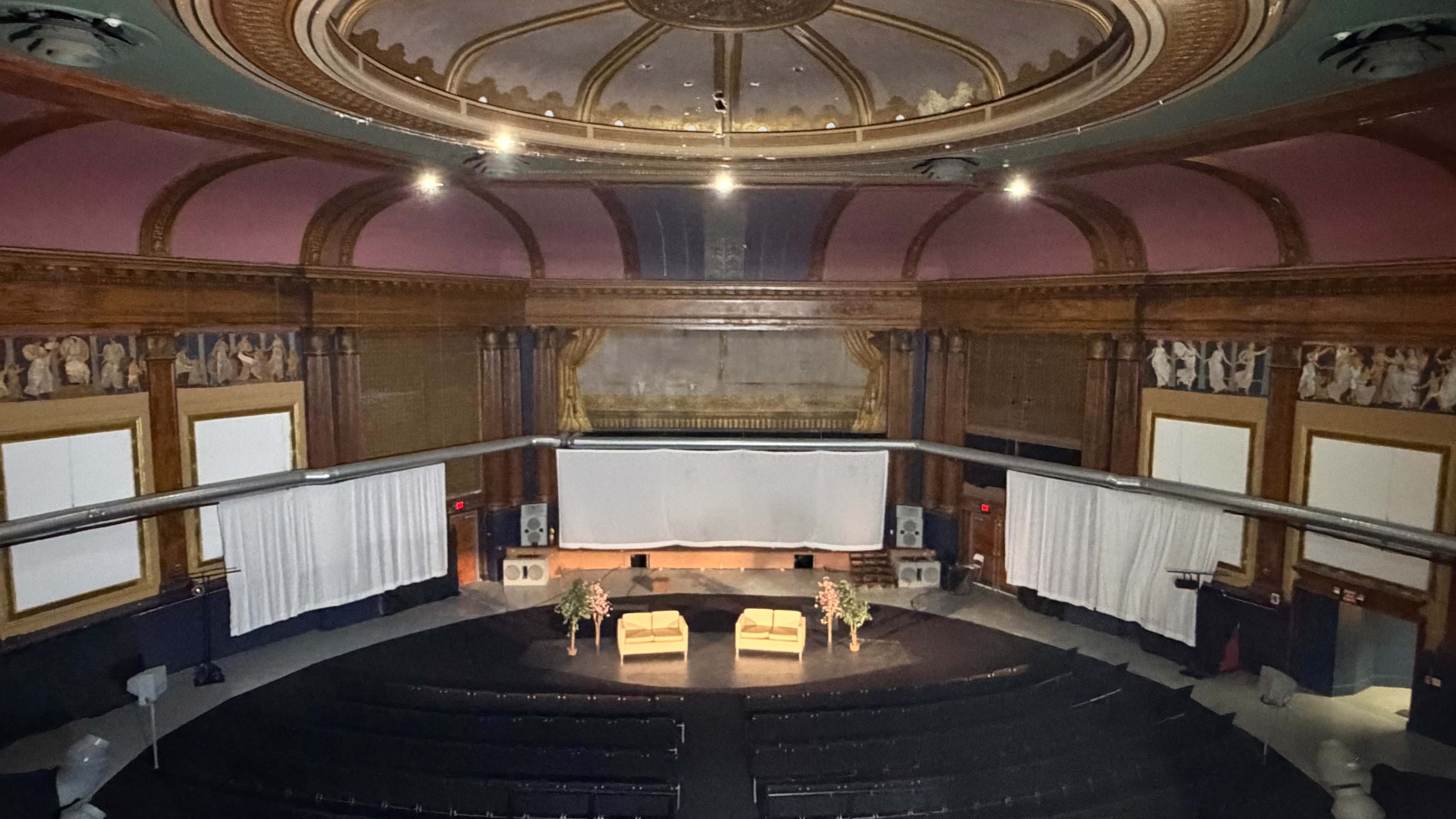 The image shows an empty auditorium with a dark stage, two chairs at the front, and decorative walls adorned with murals.