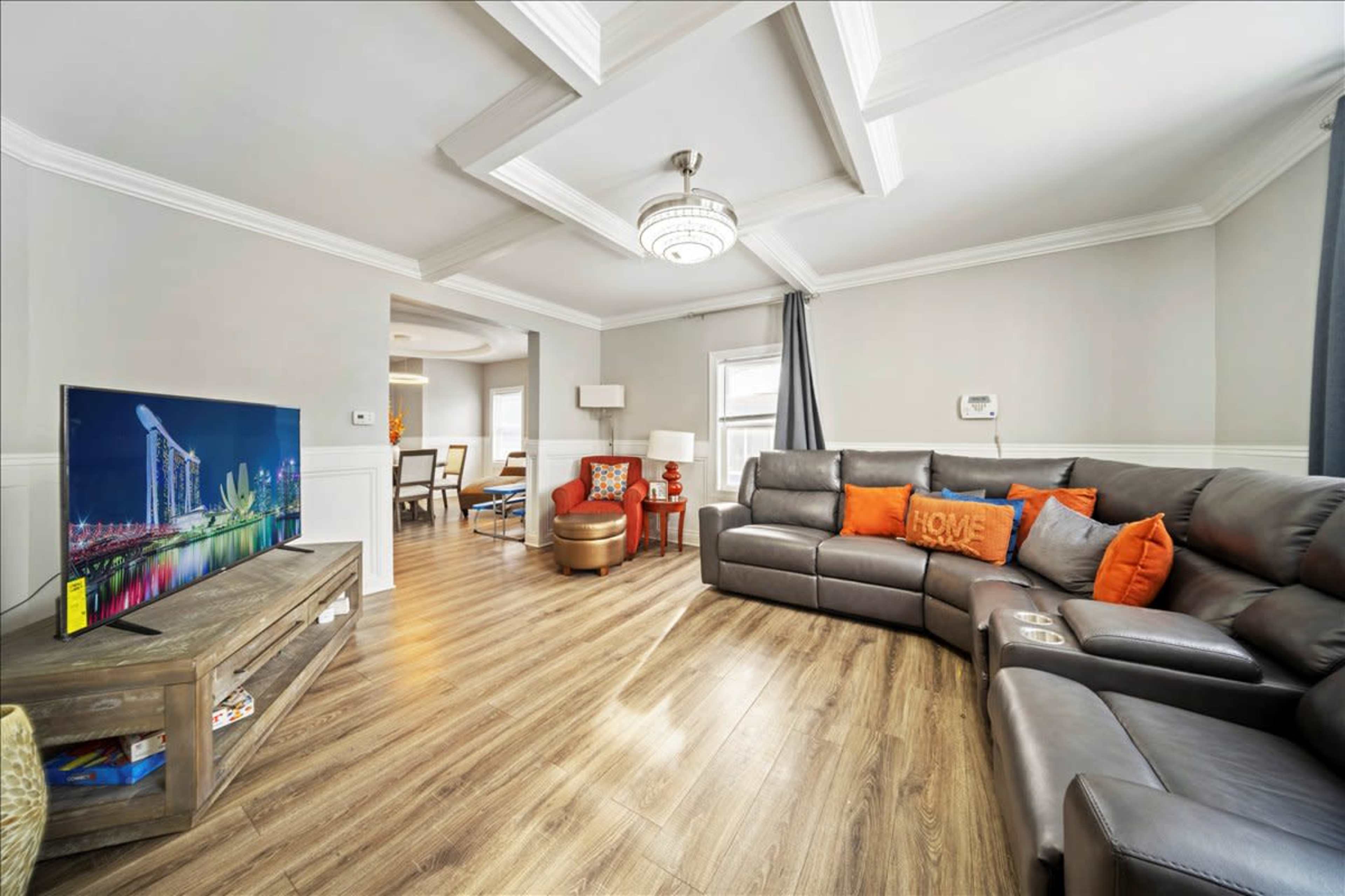 A cozy living room features a sectional couch with colorful cushions, a wooden TV stand, and a large television displaying a cityscape.