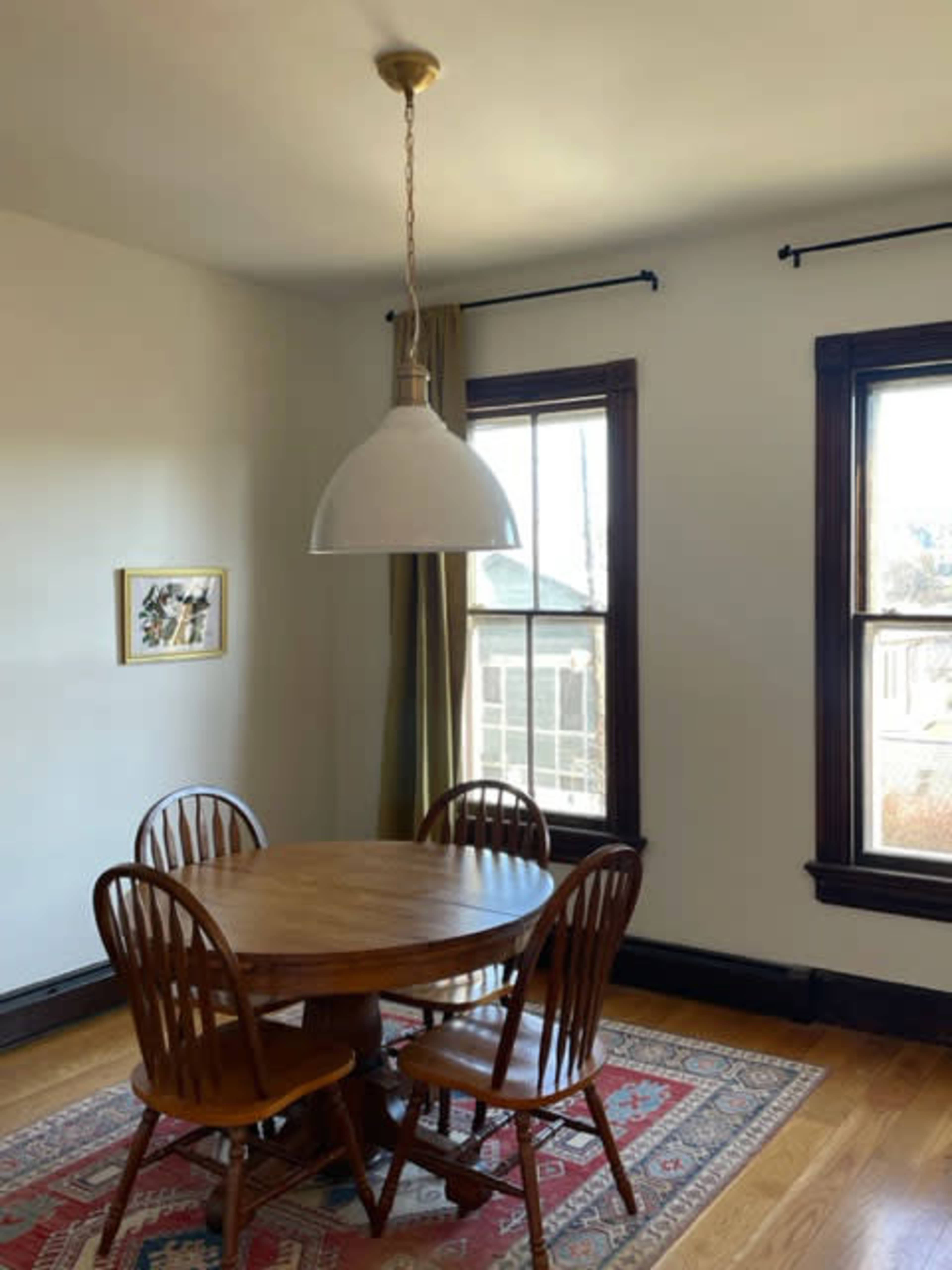 A wooden round table with four chairs is located in a room with two large windows and a hanging light fixture.