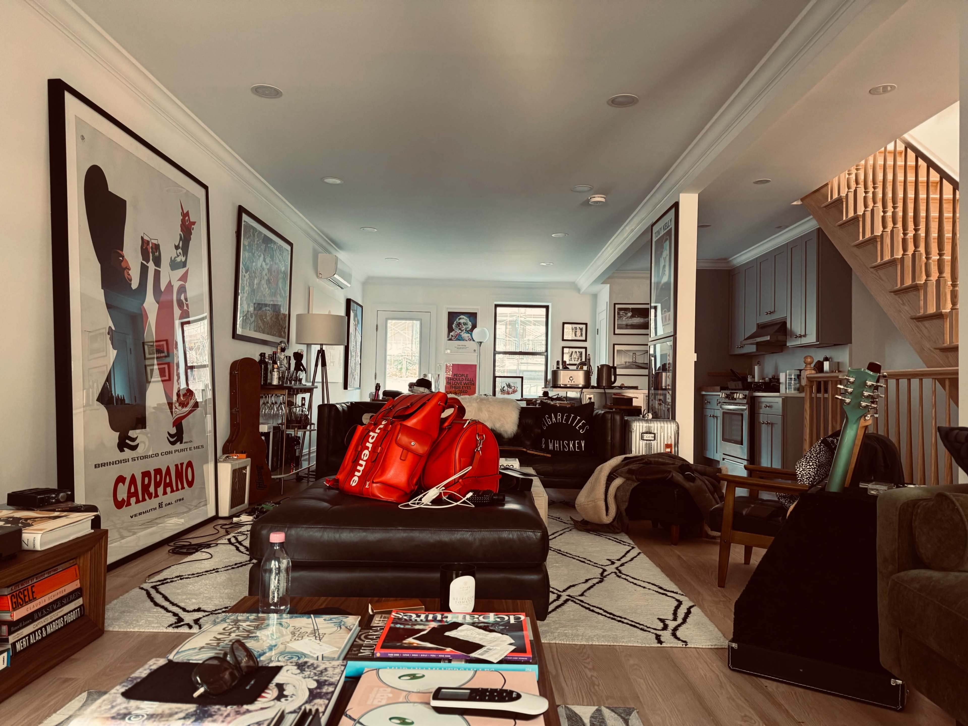 A cozy living room with a large black sofa, a red backpack, artwork on the walls, and a staircase leading to an upper level.