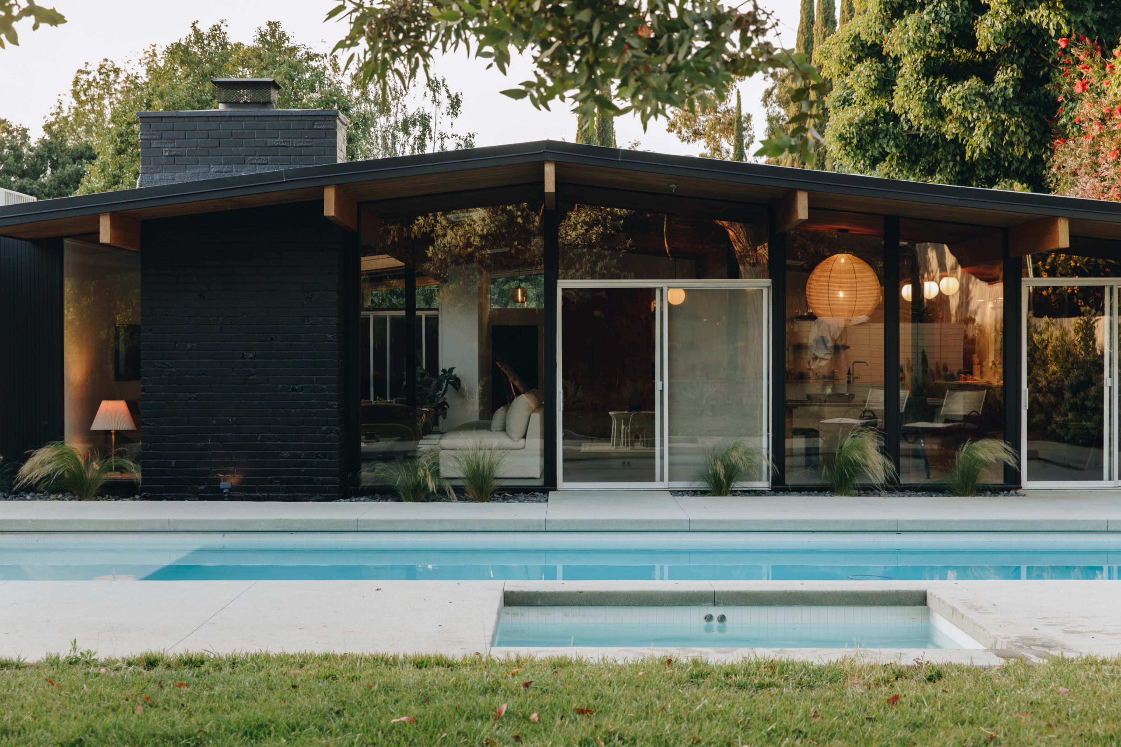 A modern, black house with large glass windows overlooks a swimming pool and is surrounded by greenery.