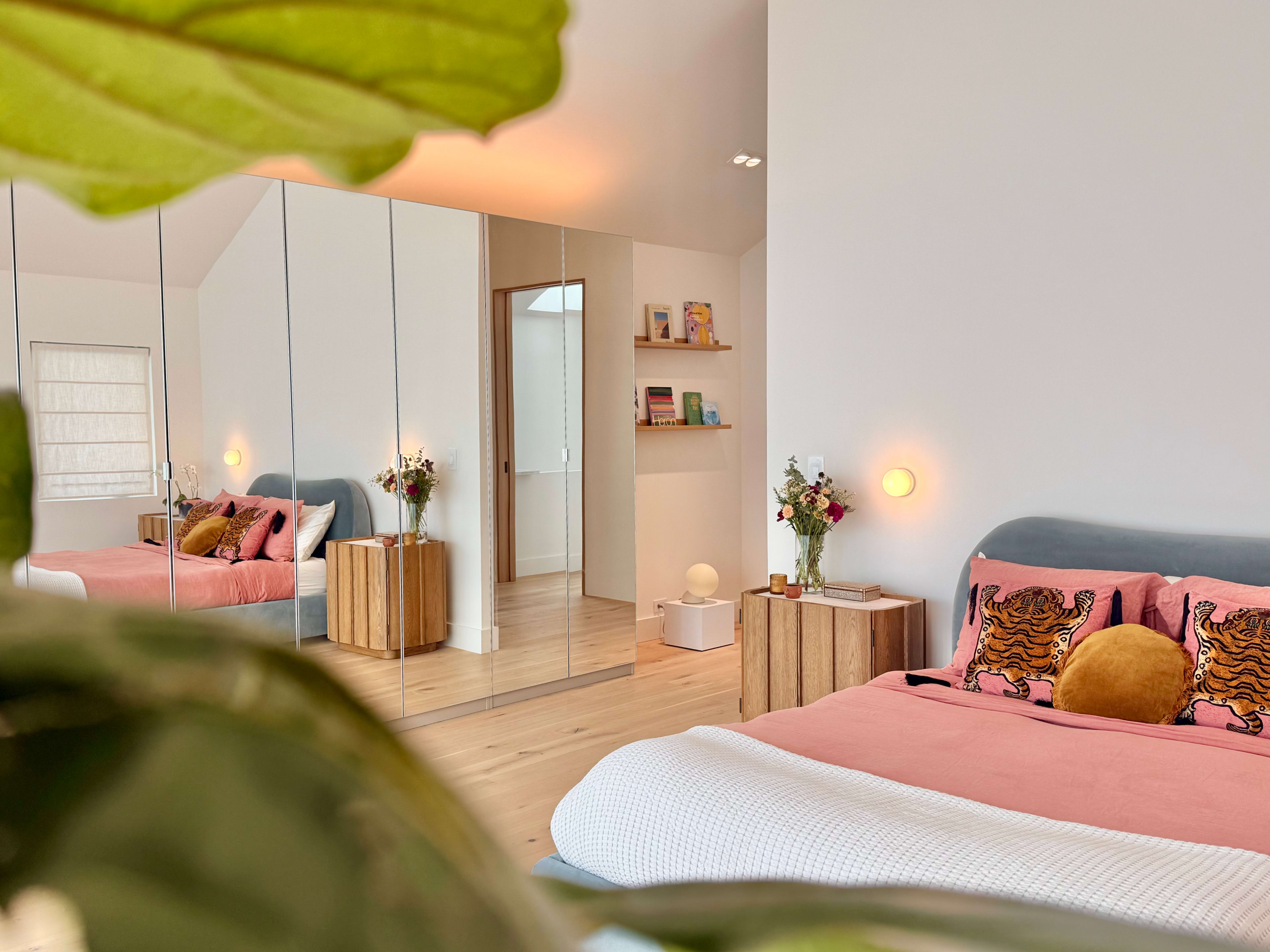 A modern bedroom features a bed with decorative pillows, a bedside table with flowers, and a mirrored wardrobe reflecting the space.