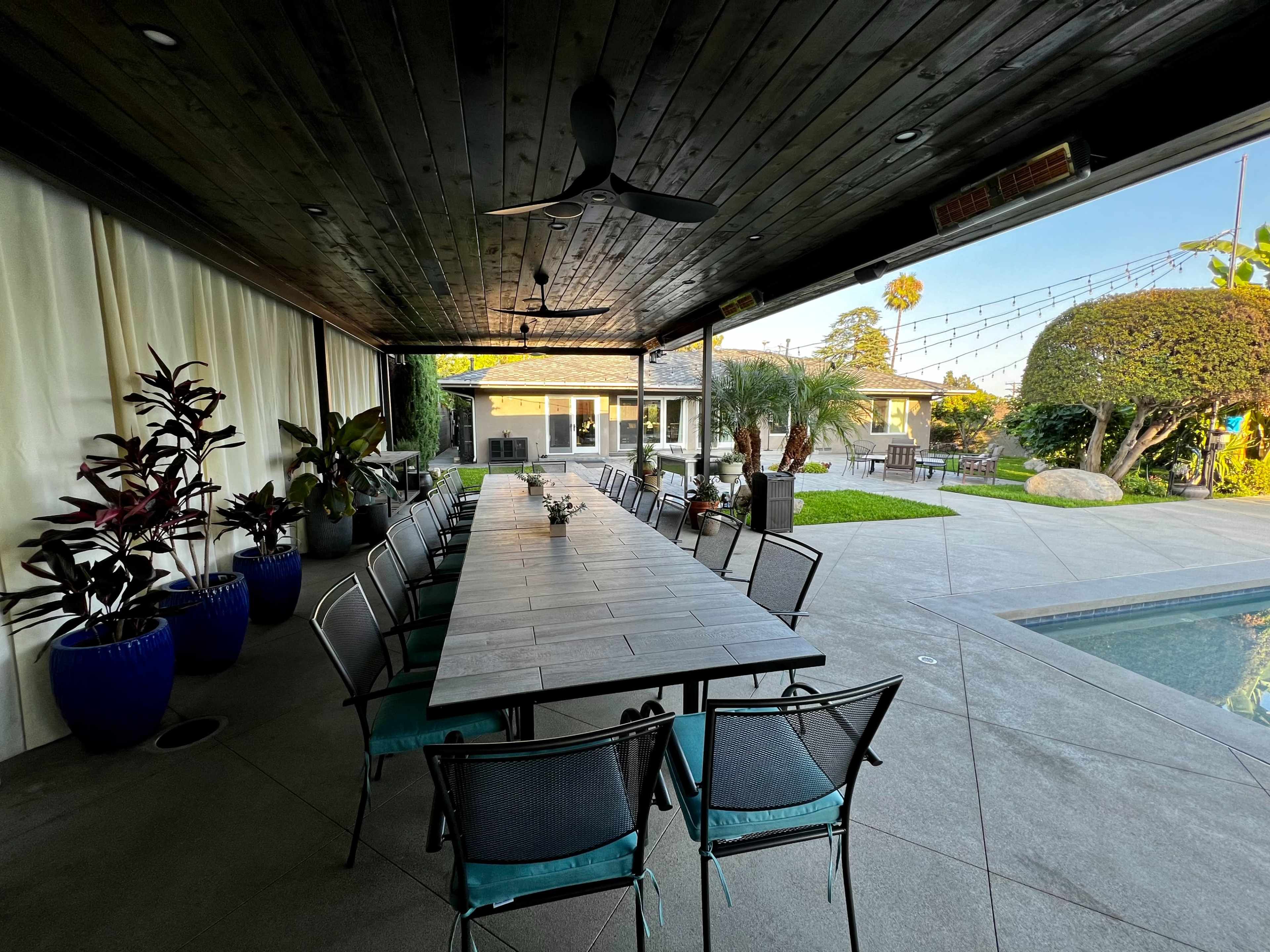 A long rectangular dining table with chairs is set up under a covered patio beside a pool, surrounded by potted plants and a well-manicured lawn.