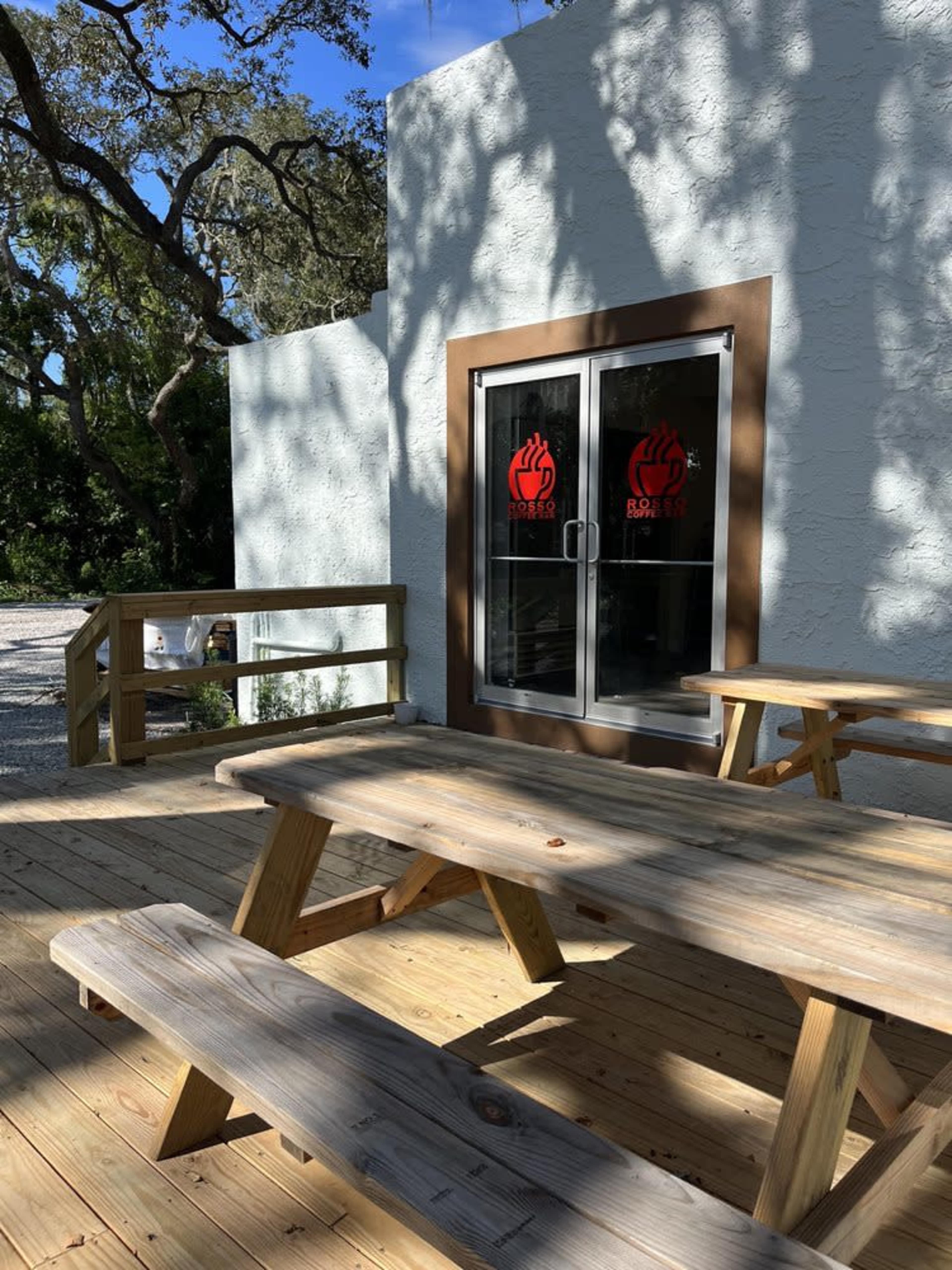 The image shows a small outdoor seating area with wooden picnic tables in front of a building featuring glass doors adorned with red designs.