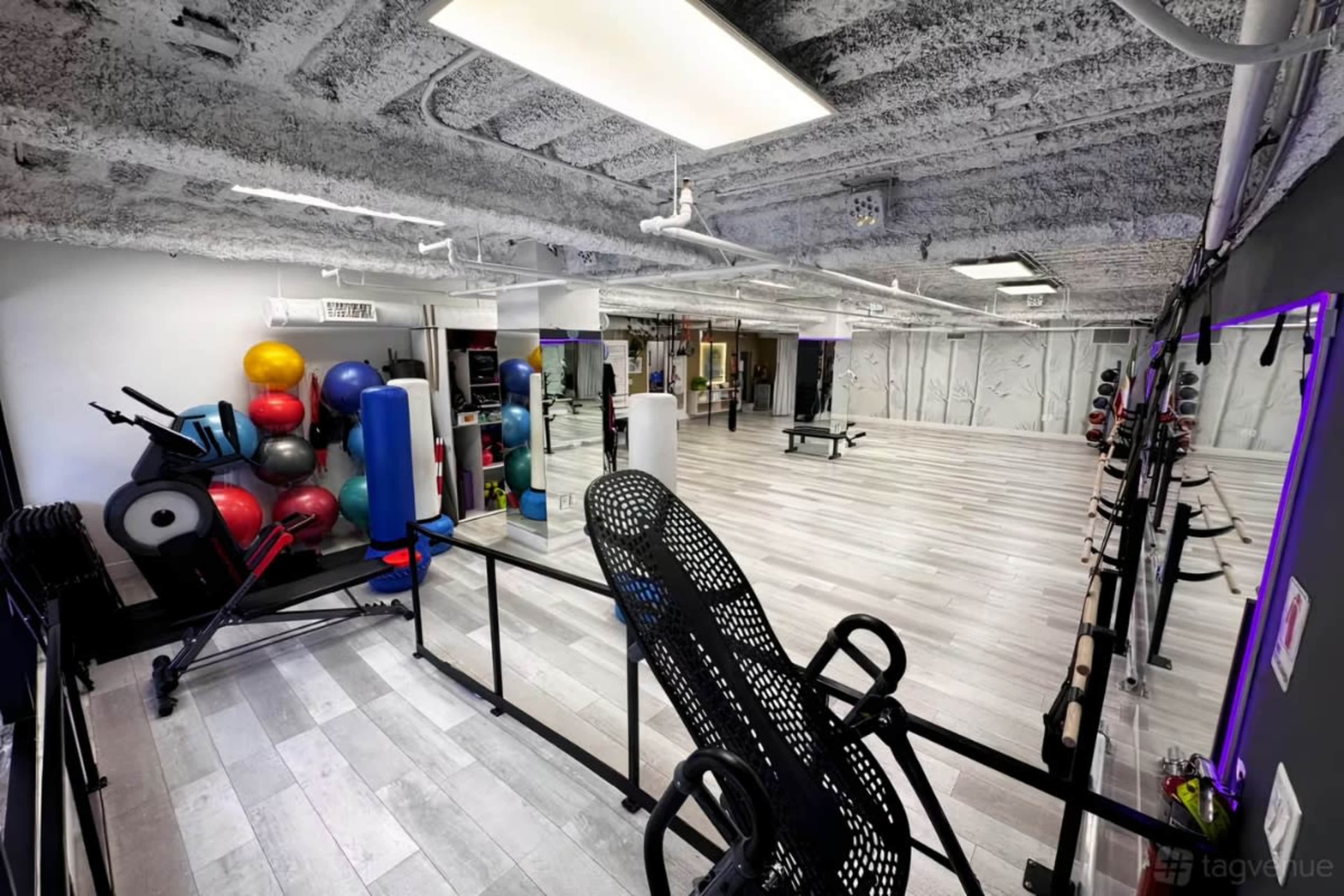 The image shows a spacious gym interior with various fitness equipment, including weights, exercise balls, and a rowing machine, against a backdrop of white walls and a textured ceiling.