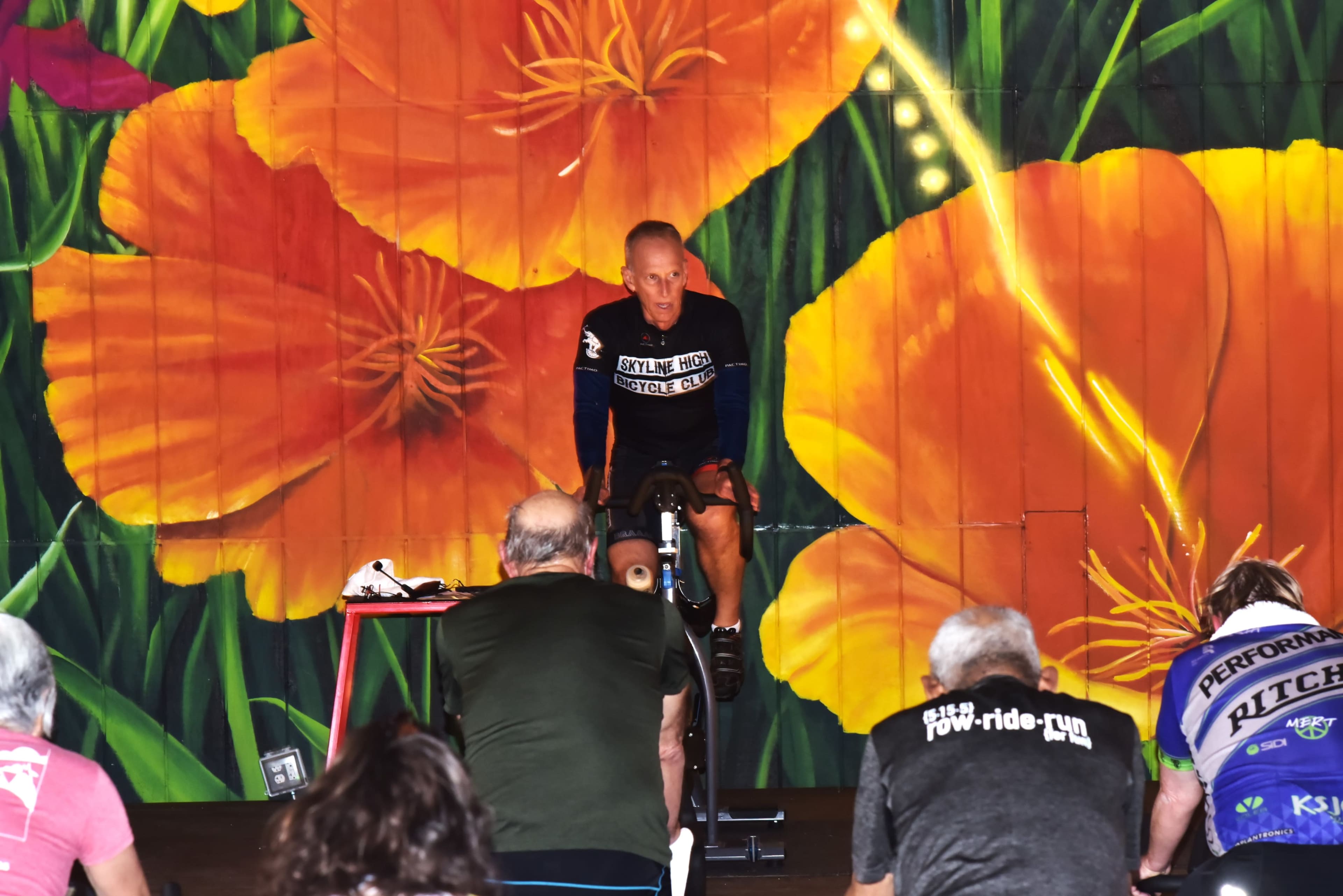 A fitness instructor leads a cycling class in front of a vibrant mural featuring large orange flowers.