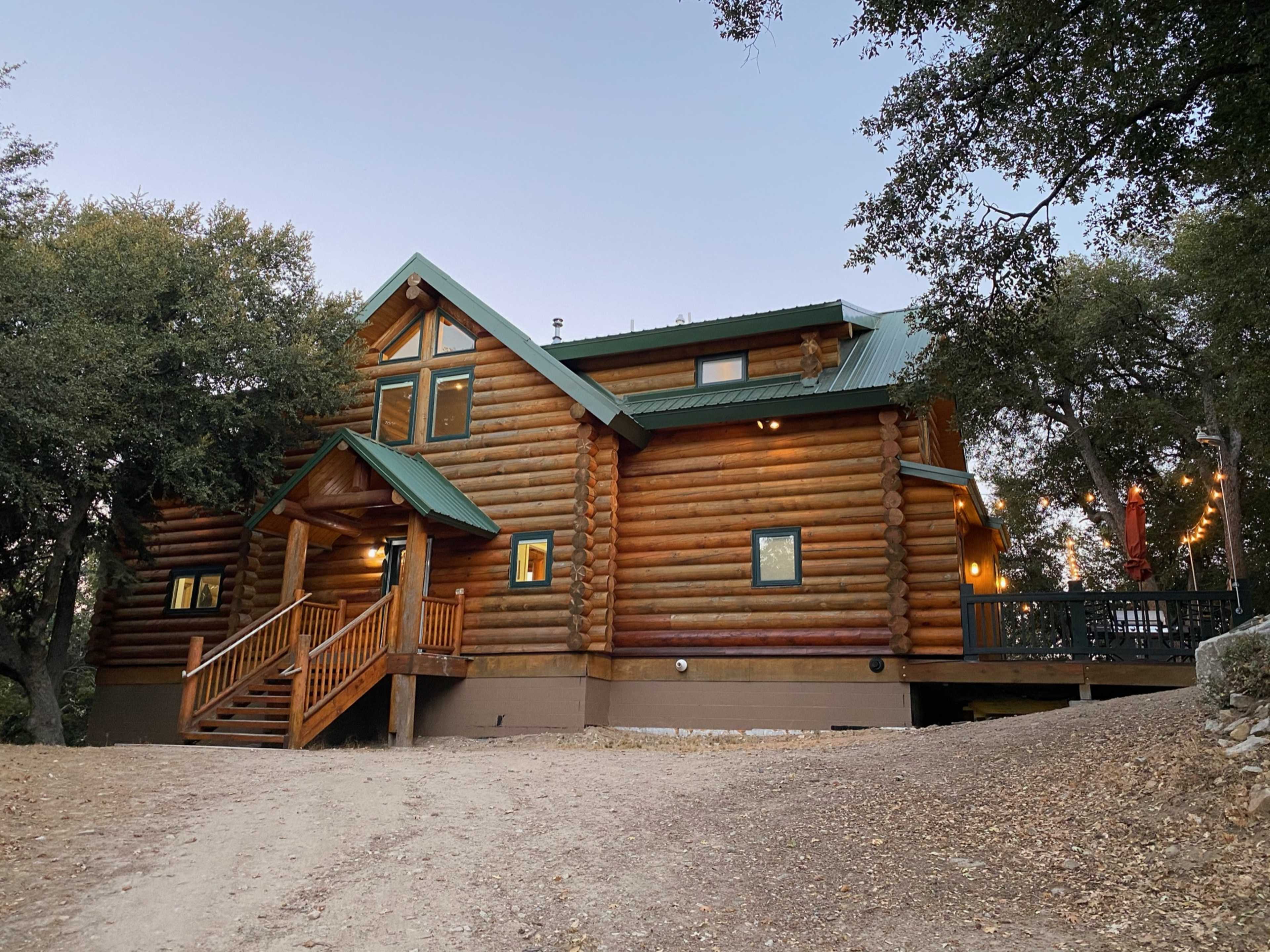 A large log cabin with a green metal roof is set on a gravel driveway, surrounded by trees.
