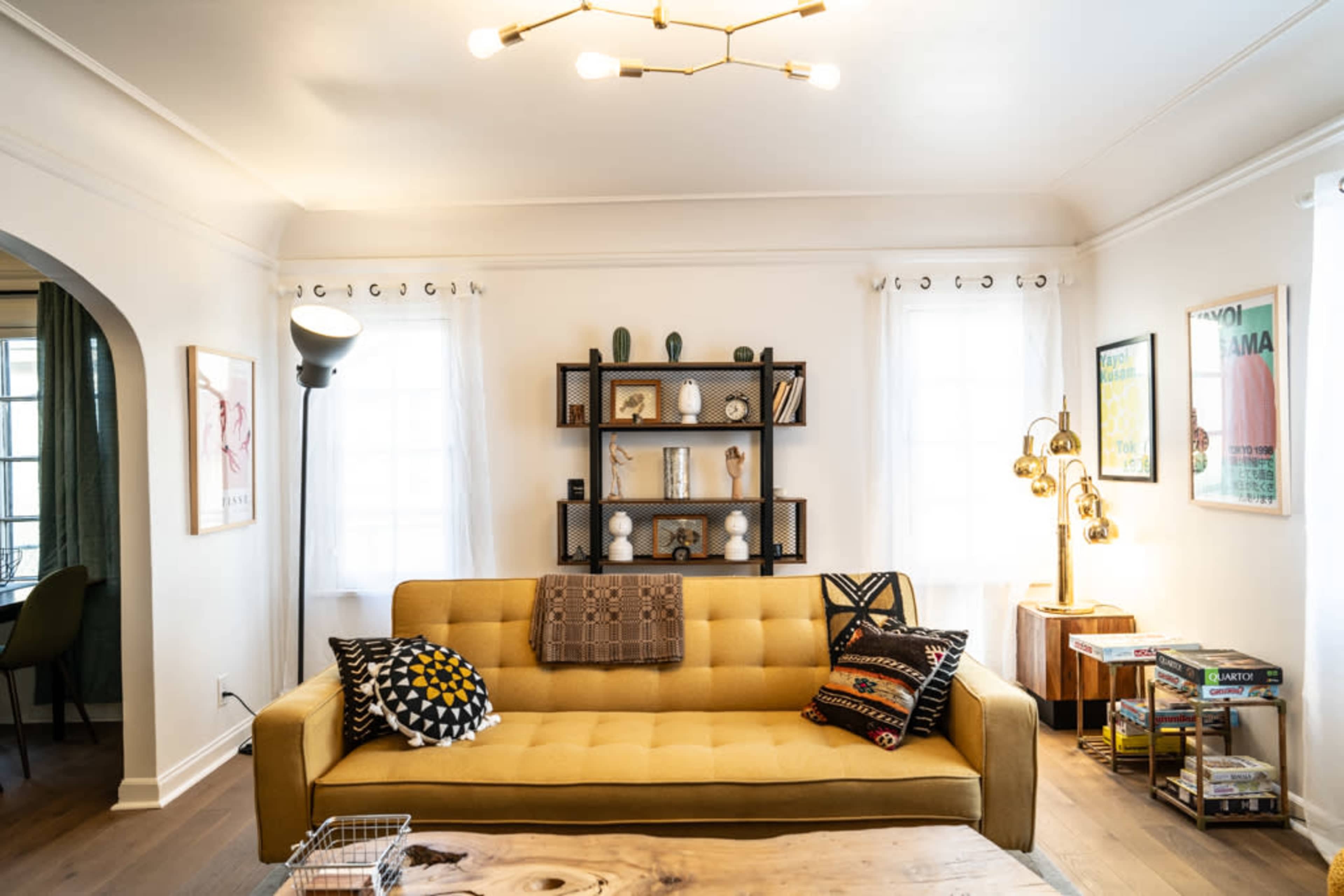 The image shows a modern living room featuring a yellow sofa with decorative pillows, a bookshelf with various artifacts, and floor lamps illuminating the space.