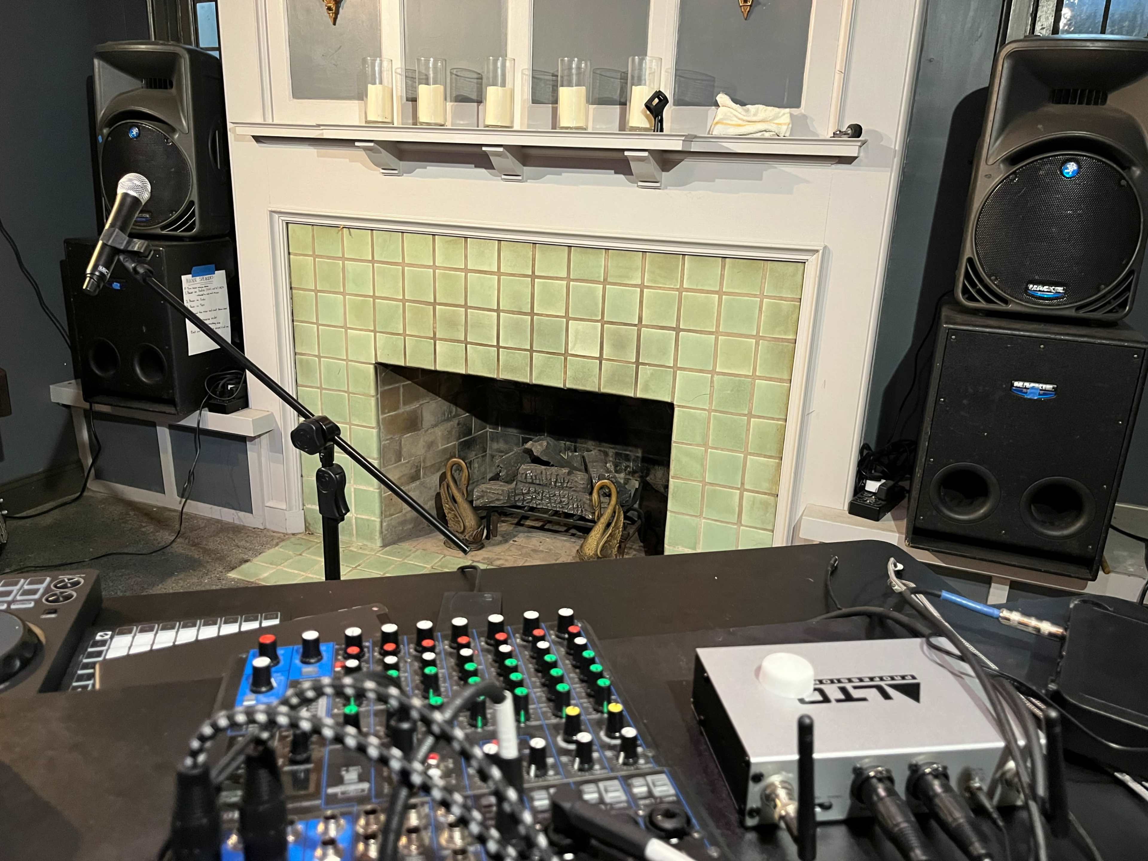 A sound mixing board in the foreground with two large speakers on either side of a brick fireplace.