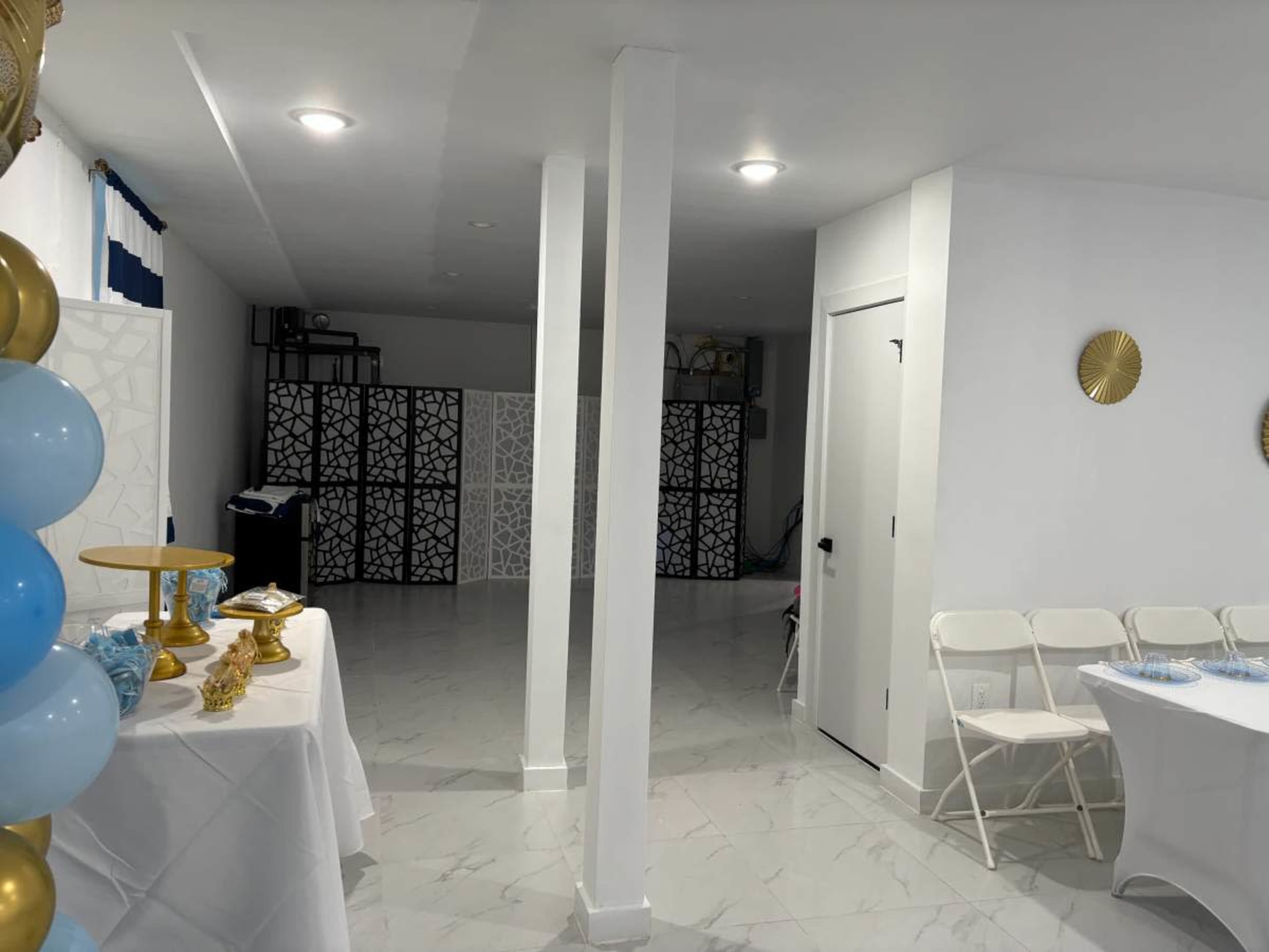 The image shows a spacious room with white walls and a tiled floor, featuring tables set with decorations and a folding partition in the background.