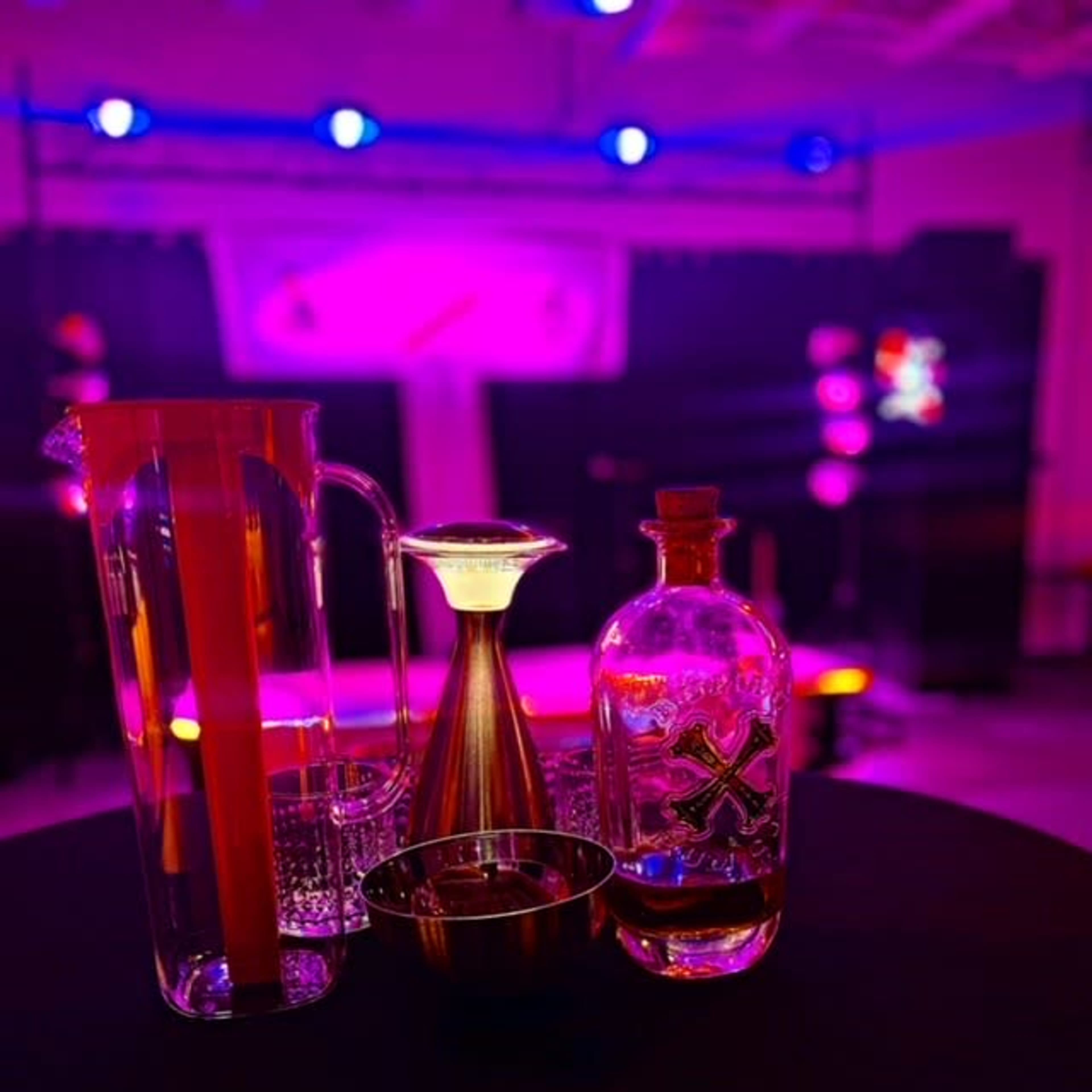 A pitcher, a metal container, and a glass bottle are arranged on a black table under purple and blue stage lighting.
