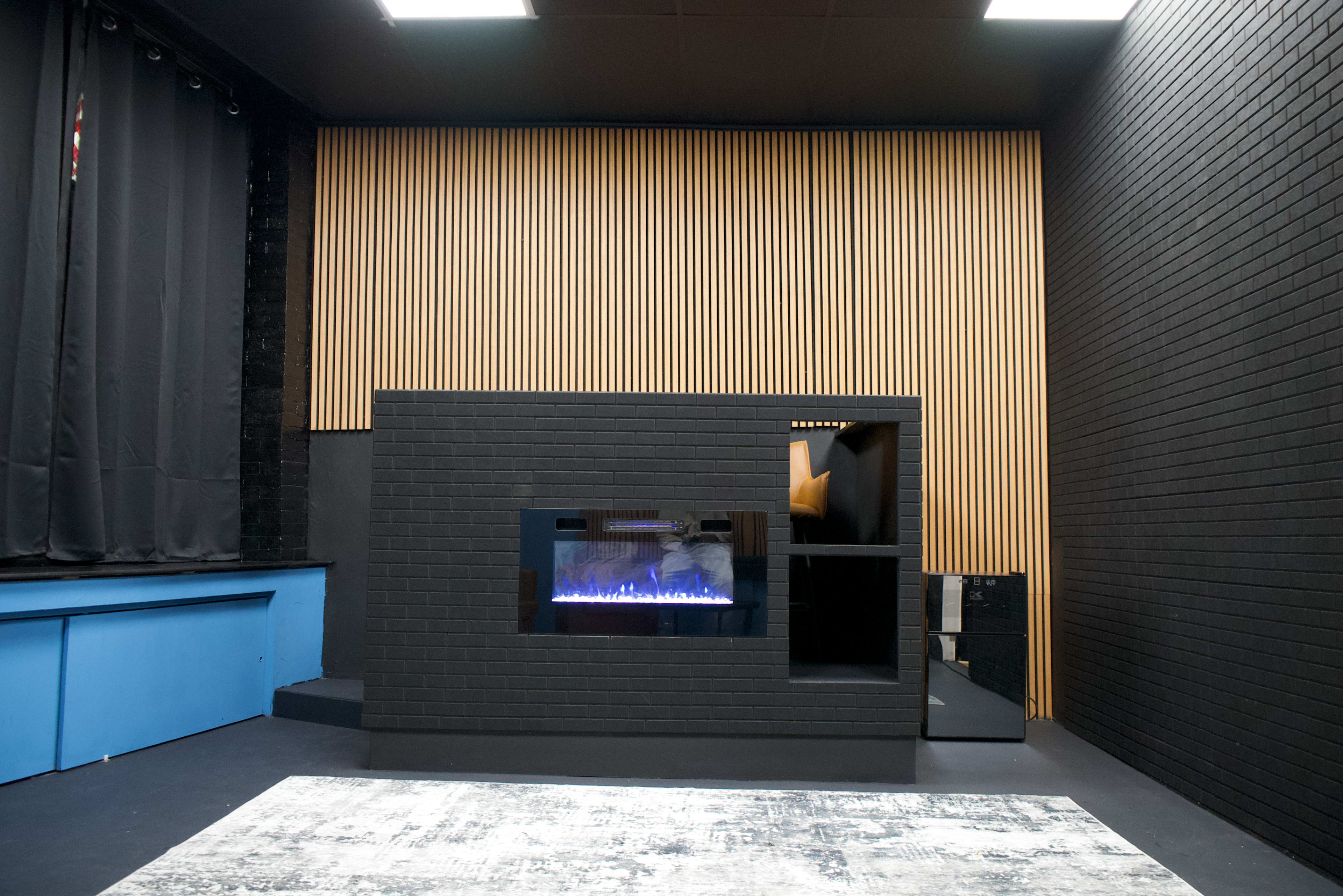 The image shows a modern interior space featuring a black brick fireplace with a blue illuminated flame, complemented by wooden paneling and a blue seating area.